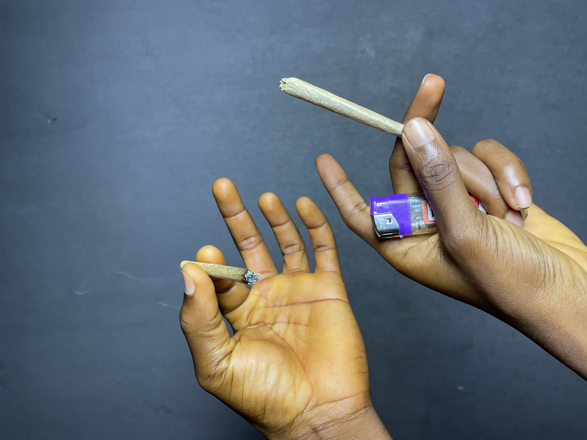 In Nkpor, Nigeria, two hands with dark skin are holding cigarettes and a lighter against a dark grey background. The hand on the left holds a lit cigarette between the thumb and index finger, with smoke rising from the tip. The palm of this hand is facing upwards and shows visible lines and slight redness. The hand on the right holds another cigarette between the thumb and index finger, and a purple and clear plastic lighter is held between the index and middle fingers of the same hand. The cigarettes appear to be hand-rolled.