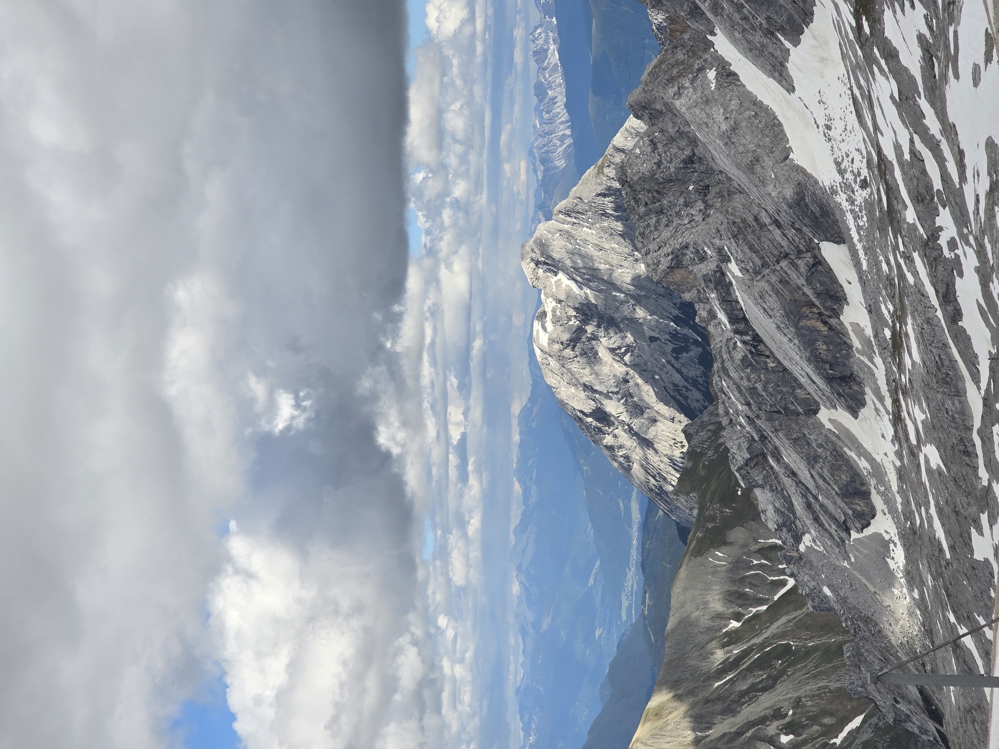 This image captures a panoramic view of the Zugspitze mountain range, the highest peak in Germany, located near Garmisch-Partenkirchen. The foreground features rugged, rocky slopes with patches of snow, indicative of a high-altitude environment. The mid-ground is dominated by a prominent, jagged mountain peak, bathed in sunlight that highlights its textures and crevices, with some snow clinging to its upper reaches. Beyond this peak, a series of layered mountain ranges recede into the distance, their blues and greens softening with atmospheric perspective. The sky above is filled with dramatic, scattered clouds, ranging from bright white cumulus to darker, heavier formations, with glimpses of clear blue sky peeking through. The overall scene conveys a sense of vastness and the raw beauty of alpine landscapes. There are no people or animals visible in the image.