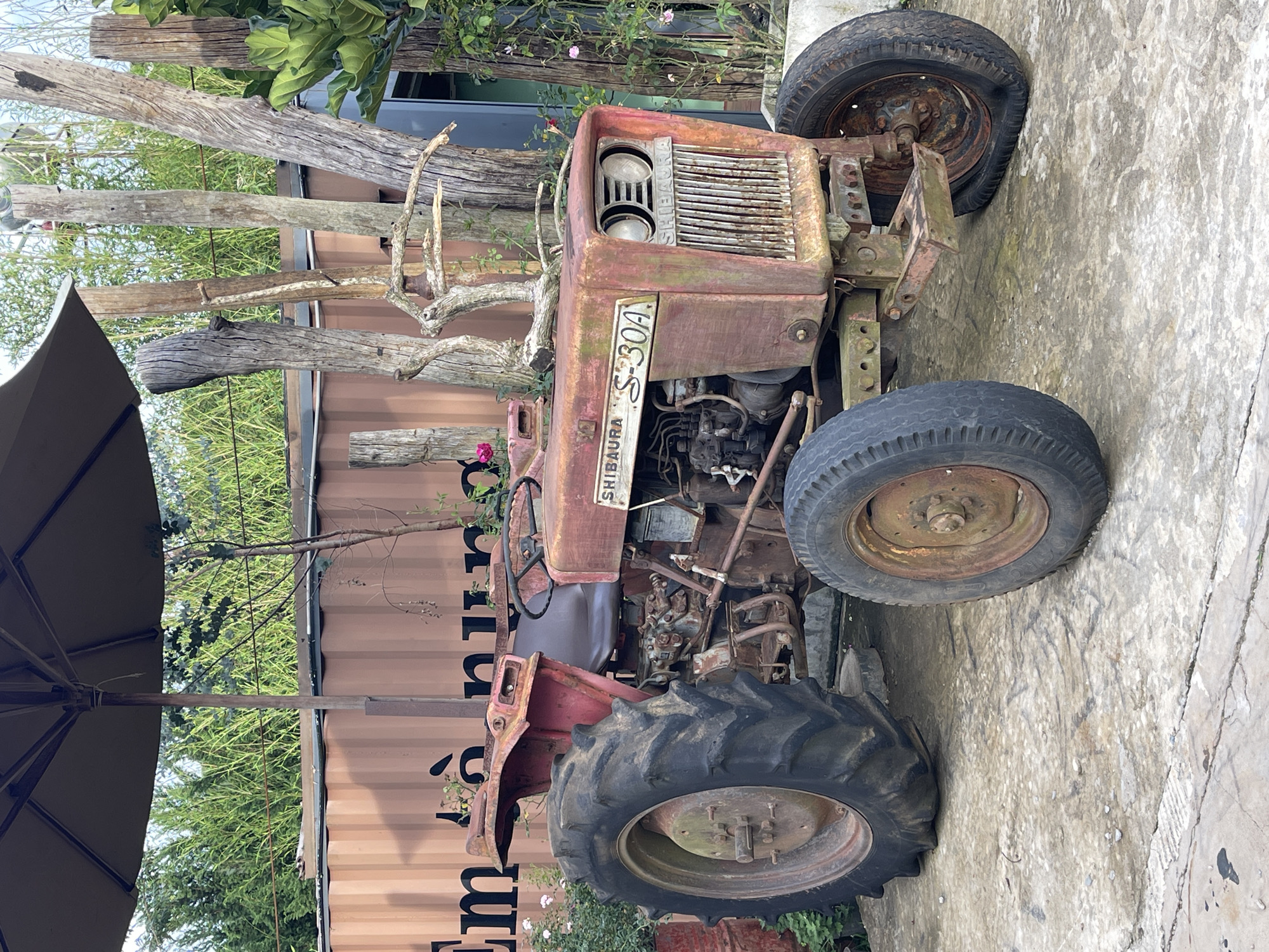 The image depicts an old, rusty Shibaura S-30A tractor on a concrete surface. The tractor has four wheels, with varying degrees of wear and tear. A wooden structure composed of branches is positioned adjacent to the tractor's front. A container and trees are visible in the background, and a sun umbrella is partially visible. The location is Thôn 5, Vietnam.