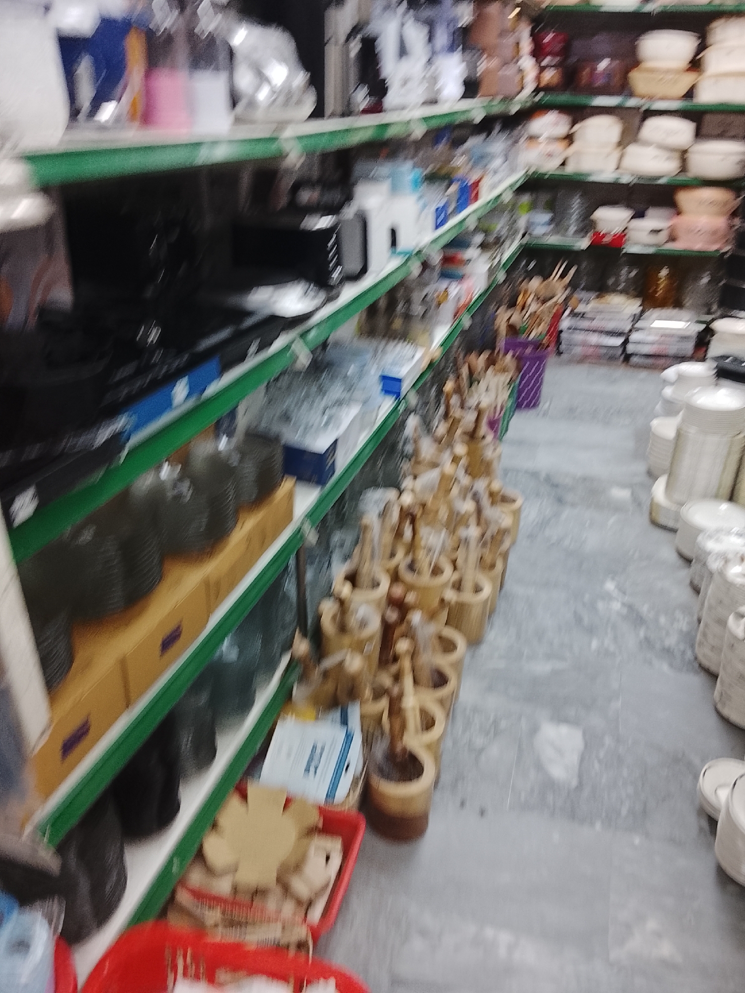 The image shows the interior of a shop in Gulgasht Colony, Pakistan. Shelves stocked with various household items such as cookware, tableware, and kitchen utensils line the walls. A large quantity of wooden mortars and pestles occupy a significant portion of the aisle. Stacks of plates and bowls are visible on the right.