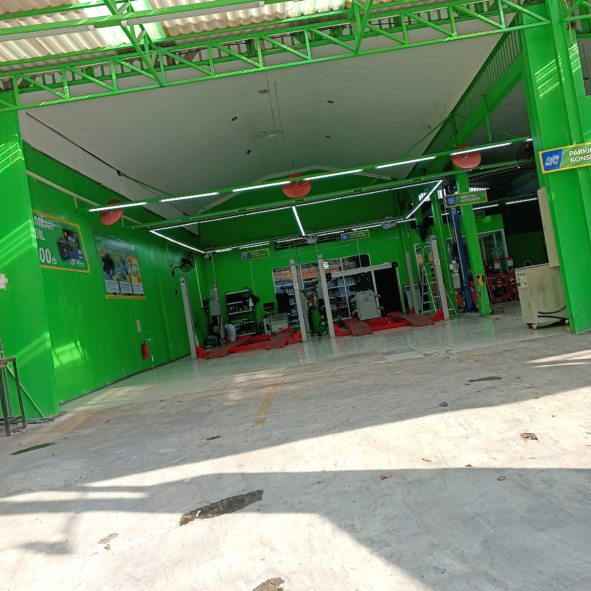 This is a wide-angle, eye-level photograph of the interior of an auto service shop in Wedoro, Indonesia, predominantly painted in bright green. The space appears to be a garage with a concrete floor. Several red car lifts are visible, positioned for vehicle maintenance. The ceiling is white, supported by a green metal truss structure, and illuminated by fluorescent strip lights. Various signs are mounted on the green walls. One sign, on the left, features an advertisement with text and an image of a battery. Another sign, further back on the left wall, indicates "AREA ISI NITROGEN" (Nitrogen Filling Area). On the right side, a yellow sign reads "PARKING KONSUMEN" (Consumer Parking). In the background, shelves stocked with bottles and containers suggest a retail or storage area for automotive fluids or parts. A tall, light-colored, wheeled appliance is positioned in the far right background. The floor has some dark stains, possibly oil or water, and scattered debris. The overall impression is a functional and organized automotive service facility.