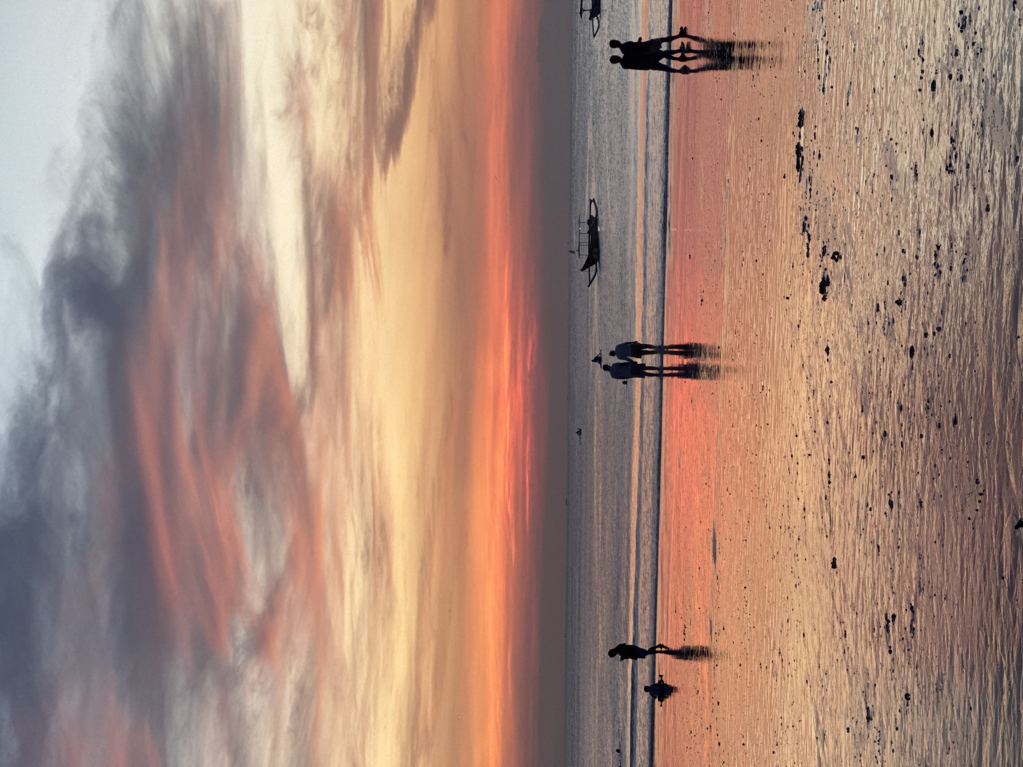 The image depicts a sunset scene on a beach in Kuta Selatan, Indonesia. The sky displays a vibrant gradient of colors, transitioning from orange and yellow near the horizon to gray at the top of the frame. Clouds are visible in the sky, adding texture to the scene.
The beach occupies the right side of the image, its wet sand reflecting the colors of the sunset. The ocean extends between the beach and the colorful sky.
Several figures are silhouetted against the sunset along the shoreline. At the top of the image, a pair of individuals stand close together, their shadows stretching along the wet sand. Another pair of people, also silhouetted, are positioned closer to the middle of the image, also casting shadows. Toward the bottom of the image, near the water's edge, are two more silhouettes of individuals. A small boat is visible, slightly off the beach.