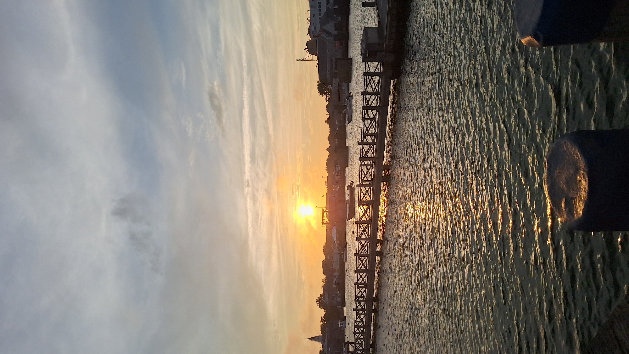 The image shows a sunset over the water in Nantucket, United States of America. A wooden boardwalk with a railing stretches across the water, leading towards a town with buildings and boats. The sun is setting on the horizon, casting a warm orange glow across the sky and reflecting on the water. In the foreground, there are two large, dark-colored bollards. The water is choppy, with ripples creating patterns on the surface. Several boats are docked or anchored in the harbor. The sky is a mix of orange, yellow, and blue, with scattered clouds.