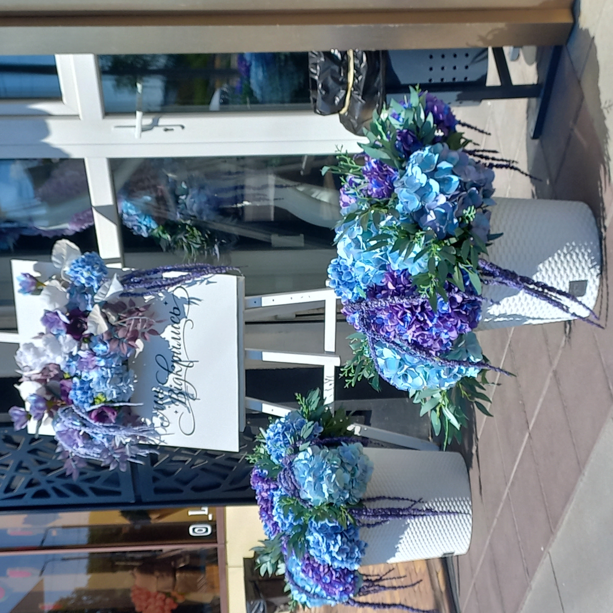 This image depicts a storefront in Kropyvnytskyi, Ukraine, adorned with a prominent floral display. The primary subjects are two large arrangements of hydrangeas and other blue and purple flowers, placed in white textured pots on the sidewalk. One pot is positioned to the left of a white easel displaying a floral arrangement and text. The text on the easel, written in a script font, appears to be "Ми відкрились" which translates to "We have opened". The storefront itself features a large glass door with white framing and a dark, ornate metal grill to the left. Reflections in the glass show interior elements of the shop. To the far left of the frame, a store window displays some merchandise, and a portion of a building facade is visible. The ground is paved with brown and grey tiles. The scene is illuminated by bright sunlight, casting shadows on the pavement. There are no people actively engaged in the scene.