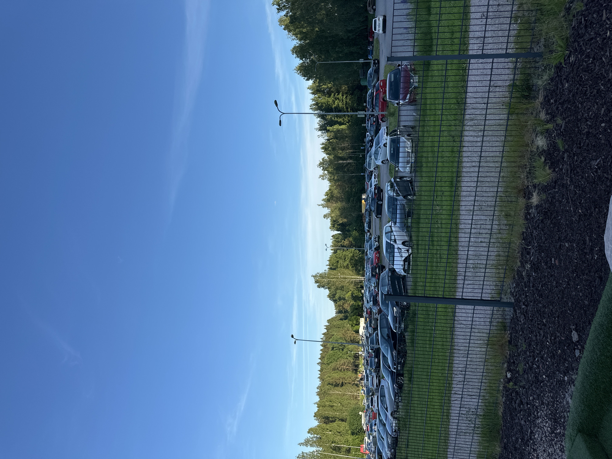 A wide shot captures an outdoor parking lot filled with numerous cars, set against a backdrop of a dense forest under a clear blue sky. The parking lot is organized, with cars parked in rows. In the foreground, a metal fence separates the parking lot from a grassy area and a path covered in gravel. The trees in the background are lush and green, indicating a temperate climate. There are no people visible in the image. The scene appears to be calm and orderly, suggesting a typical day in a suburban or semi-rural area.