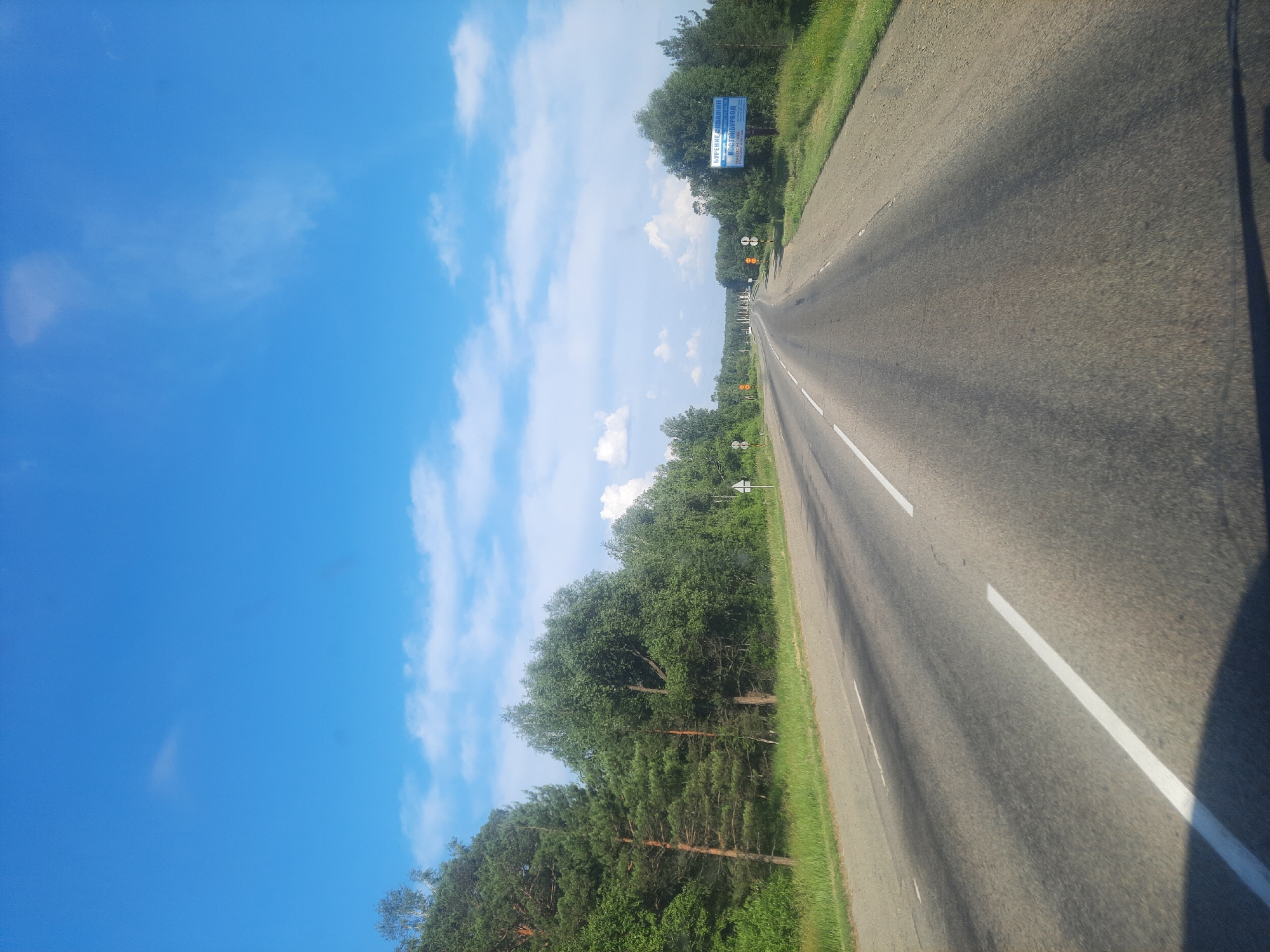 The image shows a road in the city of Баюновские Ключи, Russia. The road is asphalt, with white lane markings. On the left side of the road is a grassy area with trees, and on the right side is a sign in Russian that reads "БУРЕНИЕ СВАЛИН" and "ВОСТОЧБУРВОД". The sky is blue with some white clouds. The scene appears to be a sunny day.