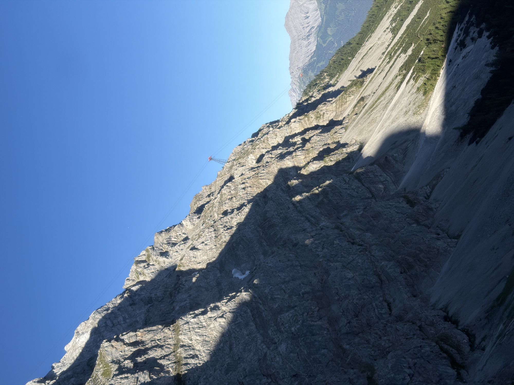 The image depicts a mountainous landscape in Grainau, Germany, likely taken from a high vantage point. The foreground is dominated by a steep, rocky mountainside casting a long shadow. The mountain is characterized by its rugged texture and striated rock formations. A thin cable stretches diagonally across the upper portion of the image, supported by pylons visible on the mountain ridges. In the distance, other mountains are visible, some with sparse vegetation and others appearing more barren. The sky is a clear, deep blue, indicating a sunny day. The overall scene conveys a sense of natural grandeur and the vastness of the alpine environment.