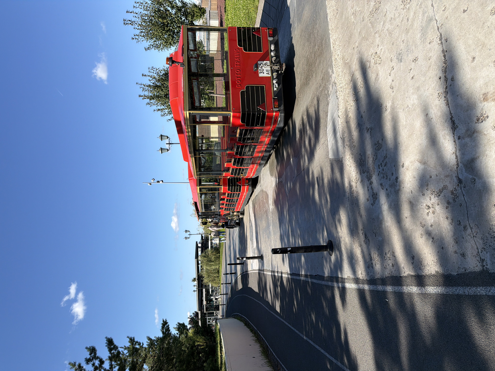 A bright red train with multiple carriages, identified by "DOTTO TRAINS" signage, is parked on a paved area under a clear blue sky with scattered white clouds. The train appears to be a tourist vehicle, with its windows and open-air seating visible. The environment suggests an outdoor recreational or tourist area, possibly a park or promenade. A paved walkway or bike path runs alongside the train, marked with white lines. Several black bollards are placed along the edge of the paved area. In the background, there are trees, a modern building with glass windows, and people walking. The scene is bathed in sunlight, casting shadows from the trees and the train onto the ground. A license plate on the back of the train reads "3045 AE 16". The presence of a train commonly found in tourist spots, coupled with the sunny weather and leisurely atmosphere, suggests it is a day of operation for the attraction.
