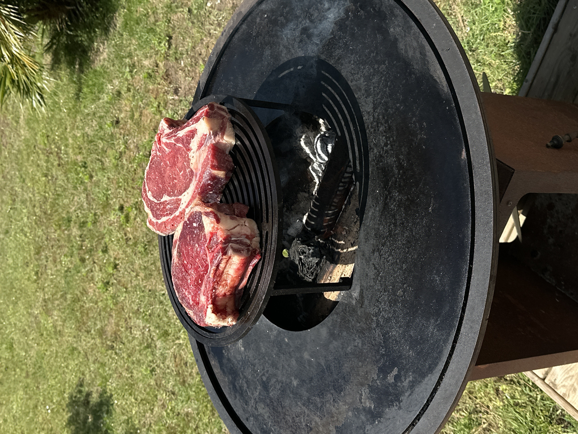 Two raw, red steaks sit on a black metal grill over smoldering wood embers. The grill is part of a larger, circular metal outdoor cooking device with a flat, table-like surface. The setting is outdoors, on a lawn with green grass and some scattered foliage. The scene appears to be in daylight. La Couarde-sur-Mer, France is not visibly identifiable in the image.