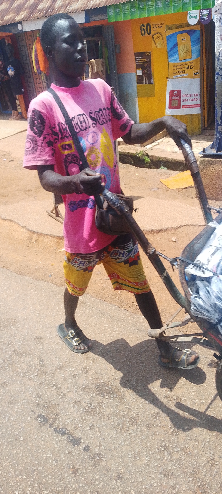 A Black male, wearing a bright pink t-shirt with a circular pattern and yellow and red patterned shorts, is pulling a wheeled cart. The cart appears to be loaded with white items, possibly textiles or produce, wrapped in plastic. He is wearing dark sandals on both feet. He has a dark brown shoulder bag across his chest. The man is walking on an asphalt road in front of a yellow and orange building that appears to be a shop or stall. The building has signage, including a large "60" and logos for MTN and Airtel. The environment suggests a street scene in a city.