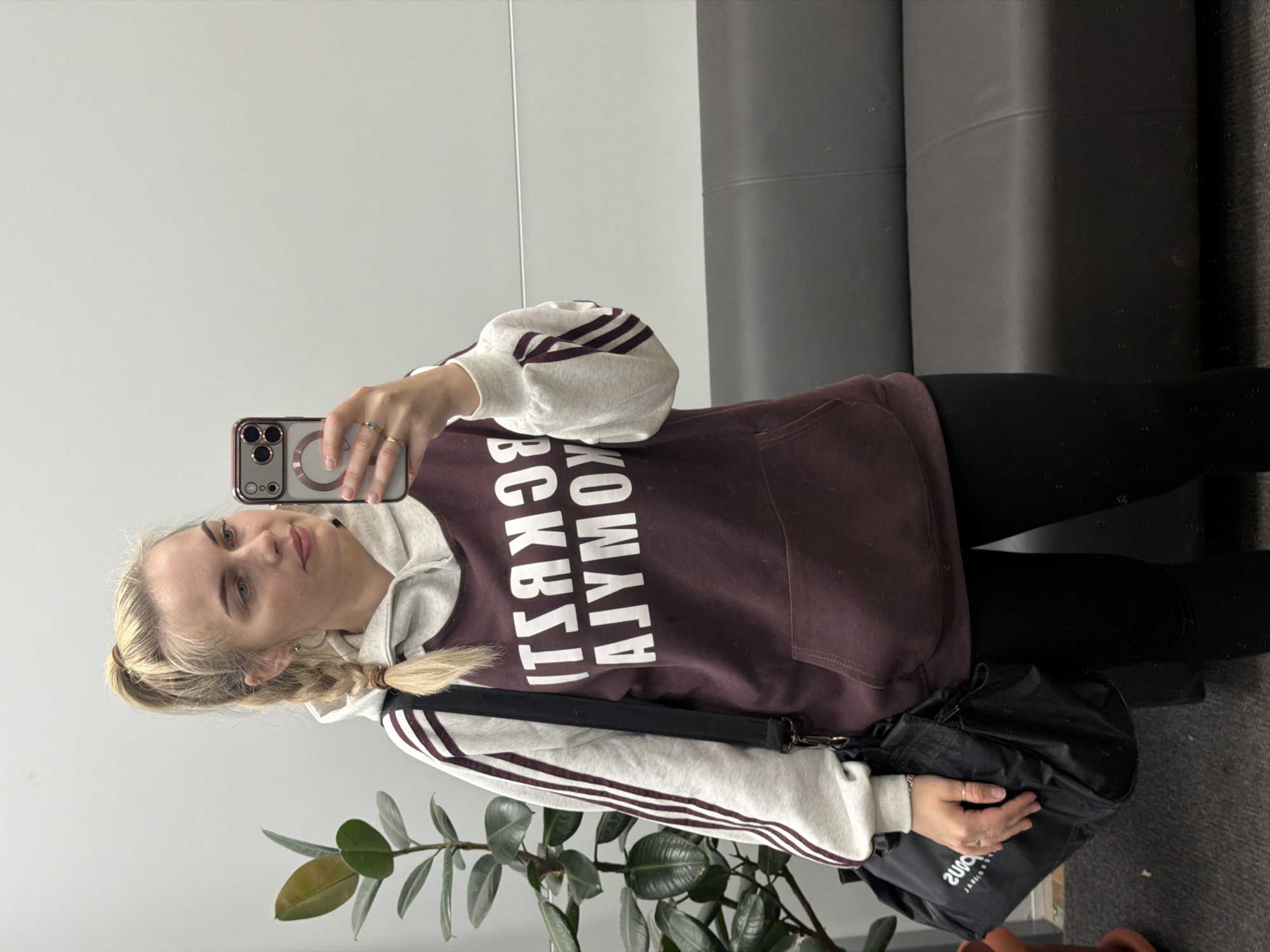 A woman stands in front of a mirrored wall and takes a selfie with a smartphone. She is wearing a burgundy hoodie with white lettering that reads "CHAIKOVSKY," a gray sweatshirt with maroon stripes on the sleeves, and black leggings. She has blonde hair styled in a braid. A black bag hangs from her shoulder. A potted plant with large, green leaves is visible in the foreground. A gray sofa is situated behind her. The location appears to be indoors. There is no explicit information to definitively confirm the location as Chaikovsky, Russia, beyond the lettering on the hoodie.