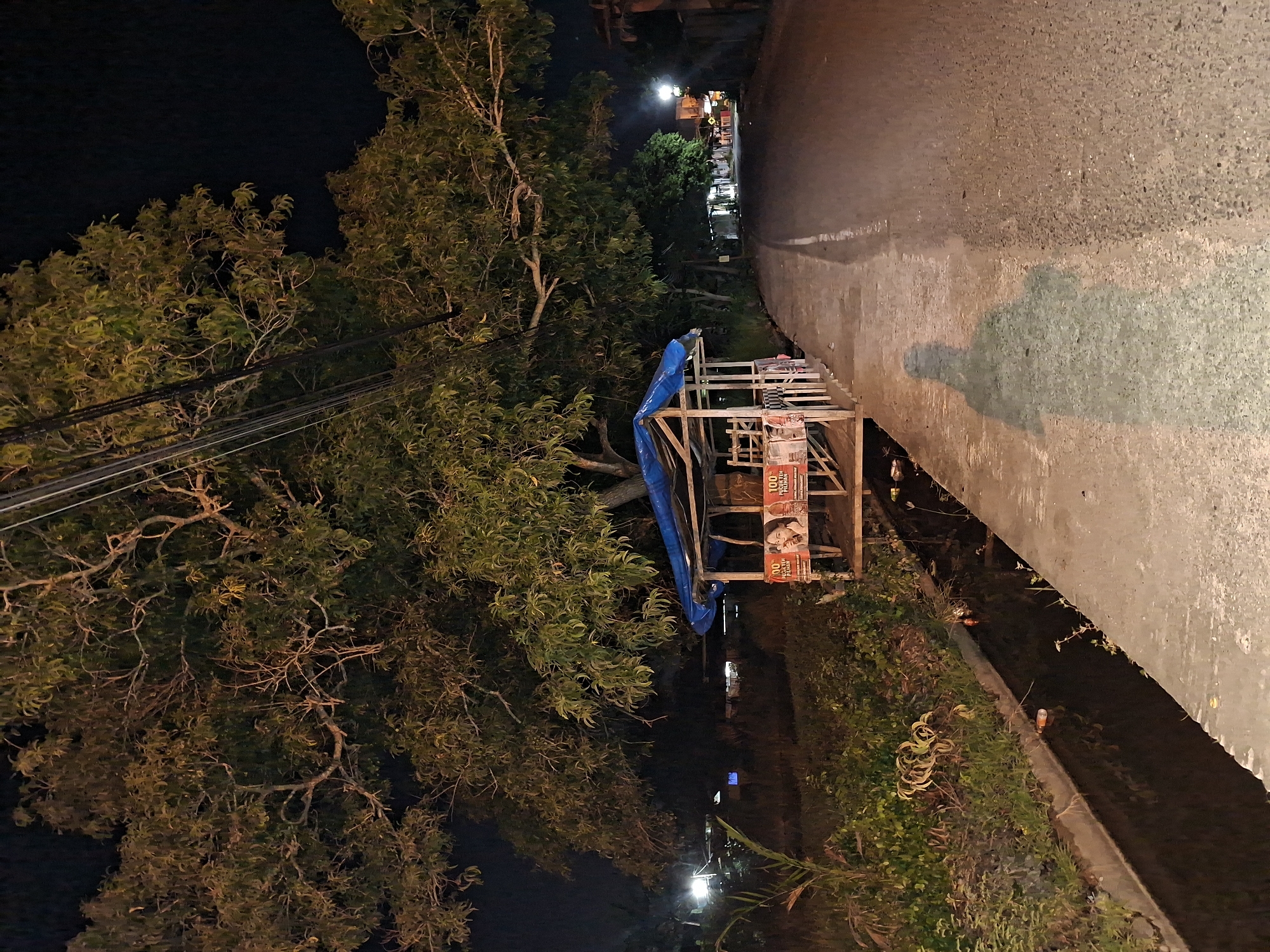 The image shows a night scene of a roadside in Lhokseumawe, Indonesia. A small wooden structure, possibly a roadside stall, sits adjacent to the road. The structure is partially covered with a blue tarp. Posters are visible on its side. A large tree with dense foliage overhangs the stall. Power lines run above the tree. The road is paved and has a visible curb. The scene is lit by streetlights in the distance. The overall environment is characterized by darkness and vegetation.