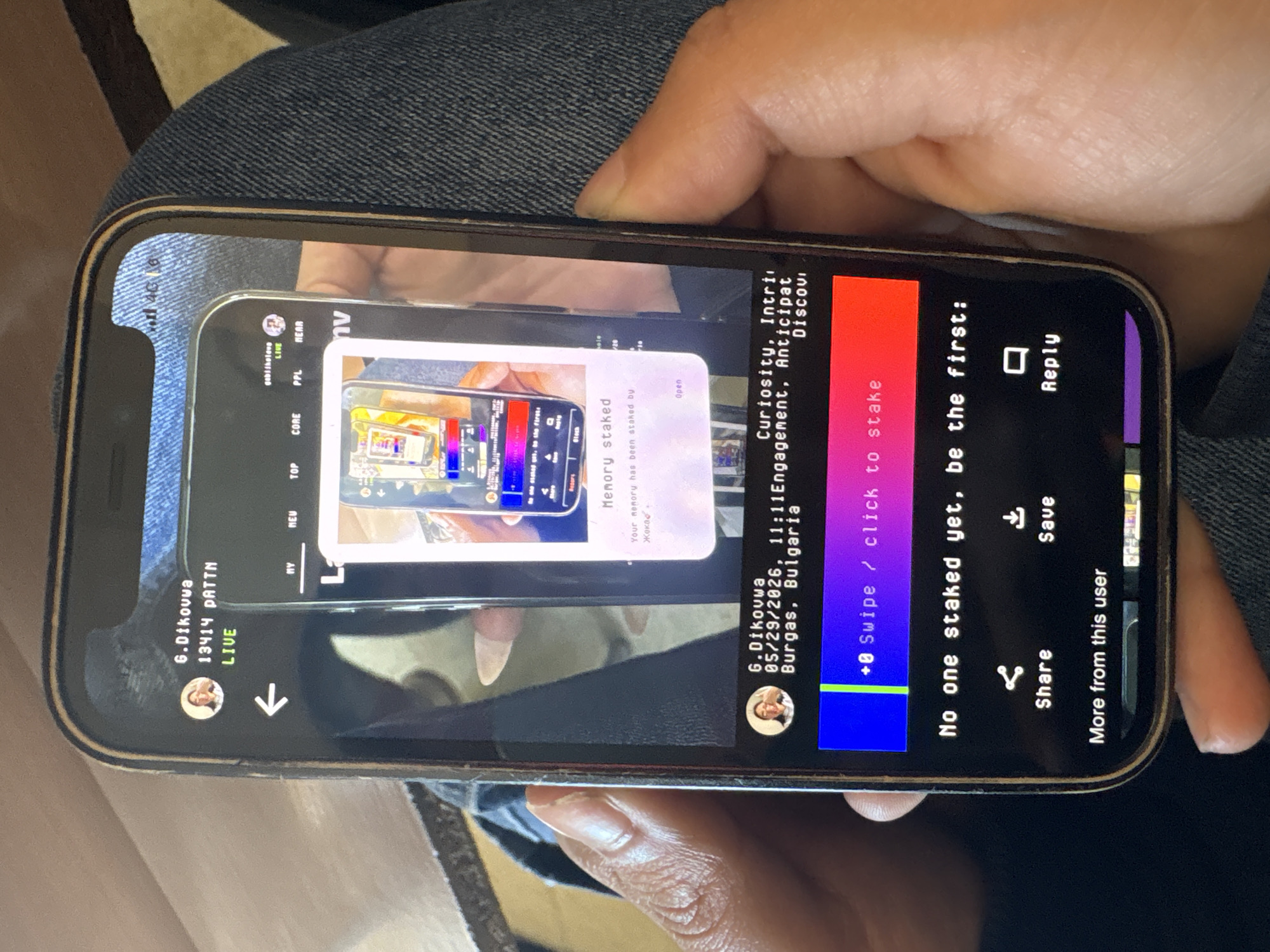 A person is holding a smartphone, displaying an image of another smartphone. The screen on the inner phone shows the text "Memory staked" and "Your memory has been staked by Жока". The outer phone displays a social media post by "6.Dikovwa" mentioning "Burgas, Bulgaria" and "05/29/2026, 11:11". It shows the text "No one staked yet, be the first:". The smartphone is being held by a person wearing denim.