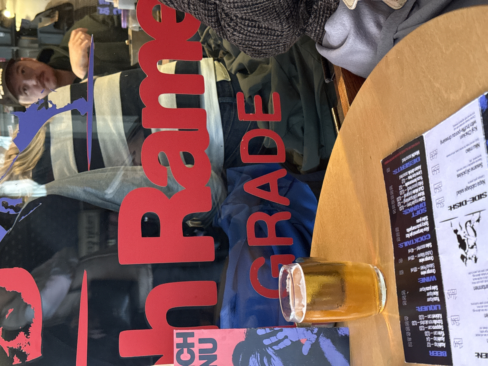 The image shows a window display in Belgrade, Serbia, with the word "Belgrade" in large red letters. A beer glass sits on a table with a menu in front of the window. The menu features drinks. The reflection of a person is visible in the window. The street scene outside the window includes clothing and other items.