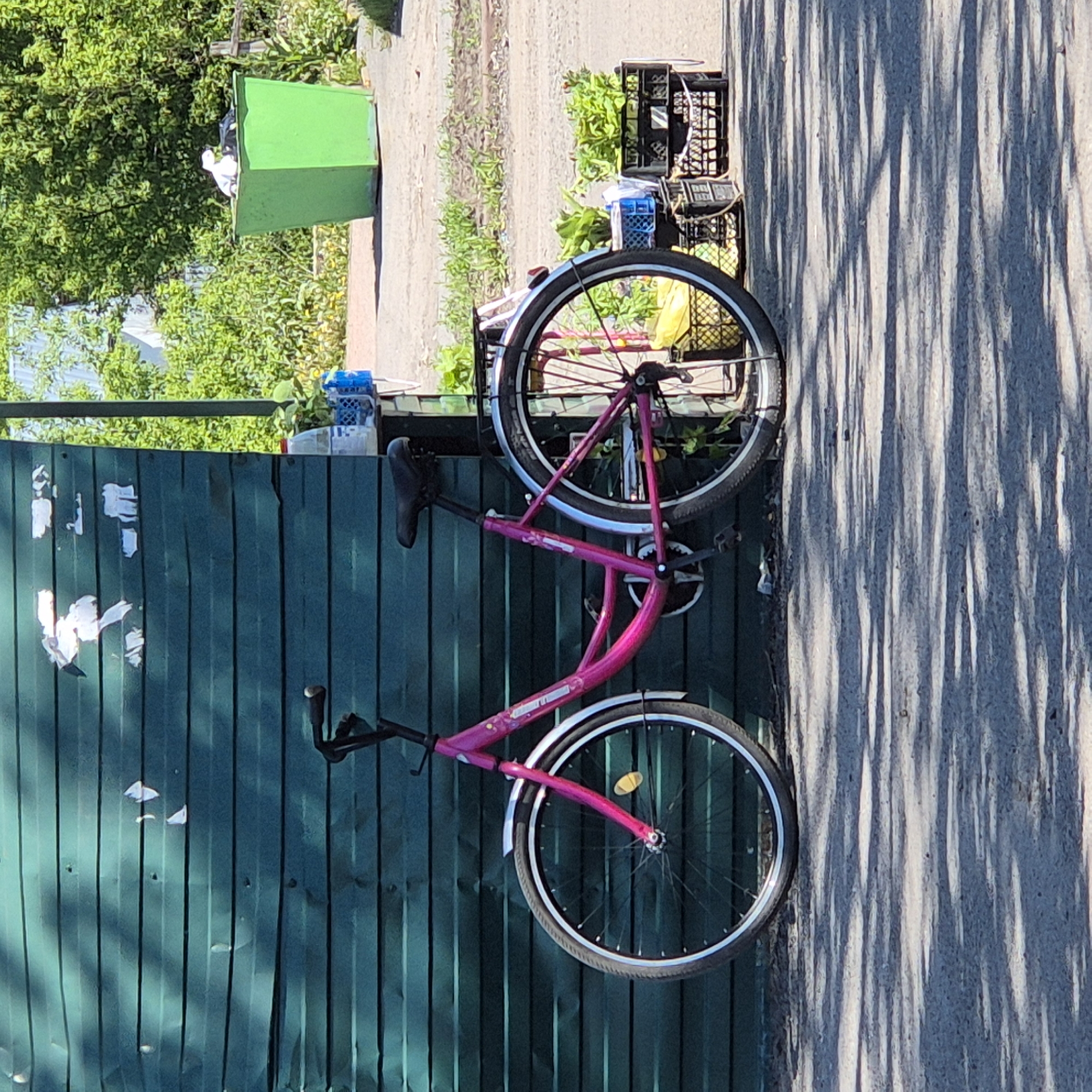 The image displays an outdoor scene in Новостройка, Russia, bathed in bright sunlight, indicated by long, distinct shadows cast across a paved surface. No people are visible.
The central subject is a vibrant pink (magenta) bicycle, positioned prominently and leaning against a dark green corrugated metal fence. The bicycle features silver fenders over both wheels, black tires, and silver-colored rims. The brand name "STELS" is faintly discernible on its frame, and a small yellow reflector is attached to the front wheel. The black seat and handlebars are also visible. The green fence, serving as a backdrop, has several pieces of white paper or tape adhered to its surface, suggesting some wear or past postings.
To the left of the bicycle, beyond the fence, a bright green dumpster or large bin is partially visible, containing what appears to be white and black waste. Lush green foliage, comprising trees and bushes, fills the background behind the fence. To the right of the bicycle, a light-colored paved area, likely a path or road, is illuminated by the sun, with strong, elongated shadows stretching across it. Near the top wheel of the bicycle, on the ground beyond the fence and near the paved area, multiple black and blue plastic crates are stacked. One black crate contains green leafy plants, and a yellow plastic bag is also visible nearby.
The length and direction of the shadows suggest the time of day is either early morning or late afternoon. The clear lighting indicates sunny weather.