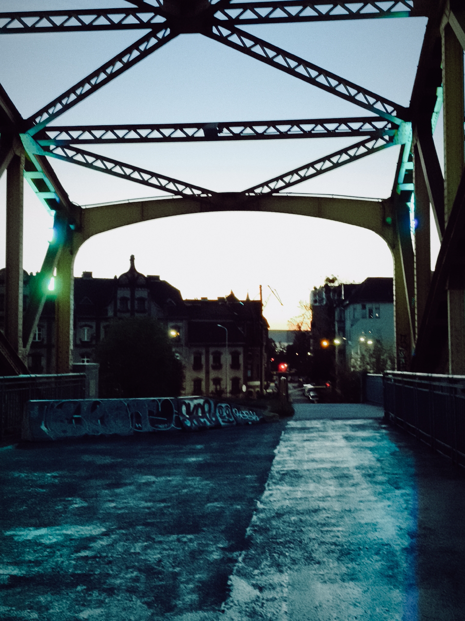 The image captures a view from a bridge in Gdańsk, Poland, likely at dusk or dawn given the low light conditions and the gradient of the sky. The foreground shows a wet, possibly reflective asphalt surface with a stark white stripe indicating a pedestrian path. To the left, a concrete barrier is covered in graffiti, rendered in a teal hue due to the lighting. The bridge's metallic structure, painted yellow and dark grey, frames the scene, with triangular patterns forming the overhead support. In the background, buildings with distinctive European architecture are visible. One building, in the center-left, has a prominent gabled roof. To the right, streetlights and traffic lights glow faintly, and the silhouette of a crane can be seen in the distance. The overall atmosphere is one of urban transition, with the decaying beauty of graffiti juxtaposed against the historical architecture and the starkness of the bridge.