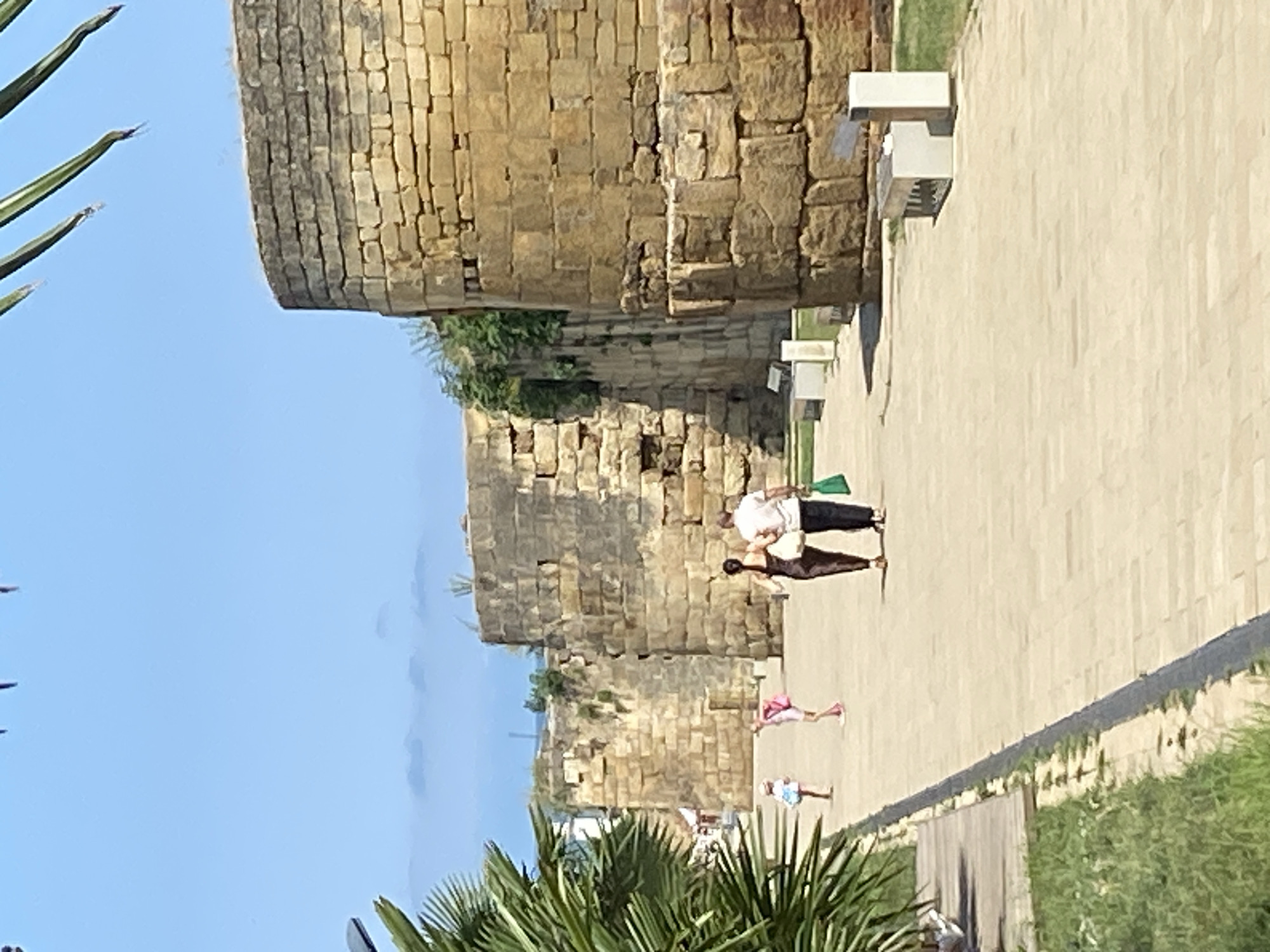 The image shows a sunny day in Derbent, Russia, focusing on the ancient stone walls of the Naryn-Kala fortress. A paved pathway runs alongside the fortress walls, bordered by green grass on the left. In the foreground, palm fronds hang from the top right, suggesting a warm climate. A couple, a man and a woman, are walking away from the camera on the pathway, the woman in a dark dress and the man in a light shirt and dark pants, carrying a green bag. Further ahead on the pathway, two young girls, one in a light pink dress and the other in a white and blue dress, are also walking away from the camera. The massive, weathered stone blocks of the fortress walls dominate the background. The sky is a clear blue with some distant, hazy clouds. Modern park benches are visible along the pathway.