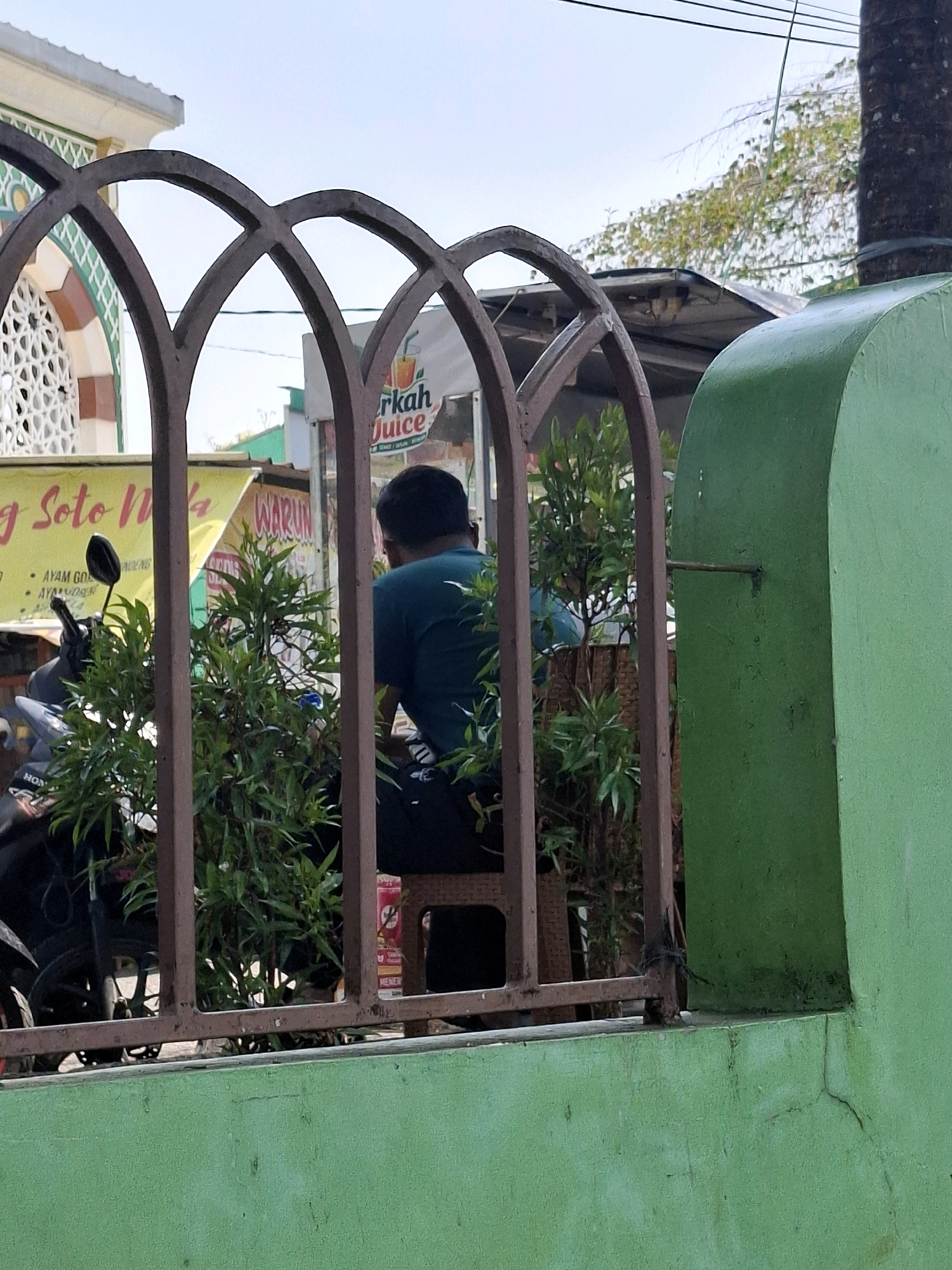 In Cianjur, Indonesia, a man wearing a teal t-shirt and black pants sits on a chair behind a brown metal fence. He is facing away from the camera and looking towards a stall. In front of him are lush green plants. Behind the plants, a motorcycle is parked. In the background, a building with a white roof and decorative green and white patterns is visible, possibly a mosque. A yellow banner with red text and a picture of a glass is also visible, suggesting a food stall. The scene is outdoors under a bright sky.