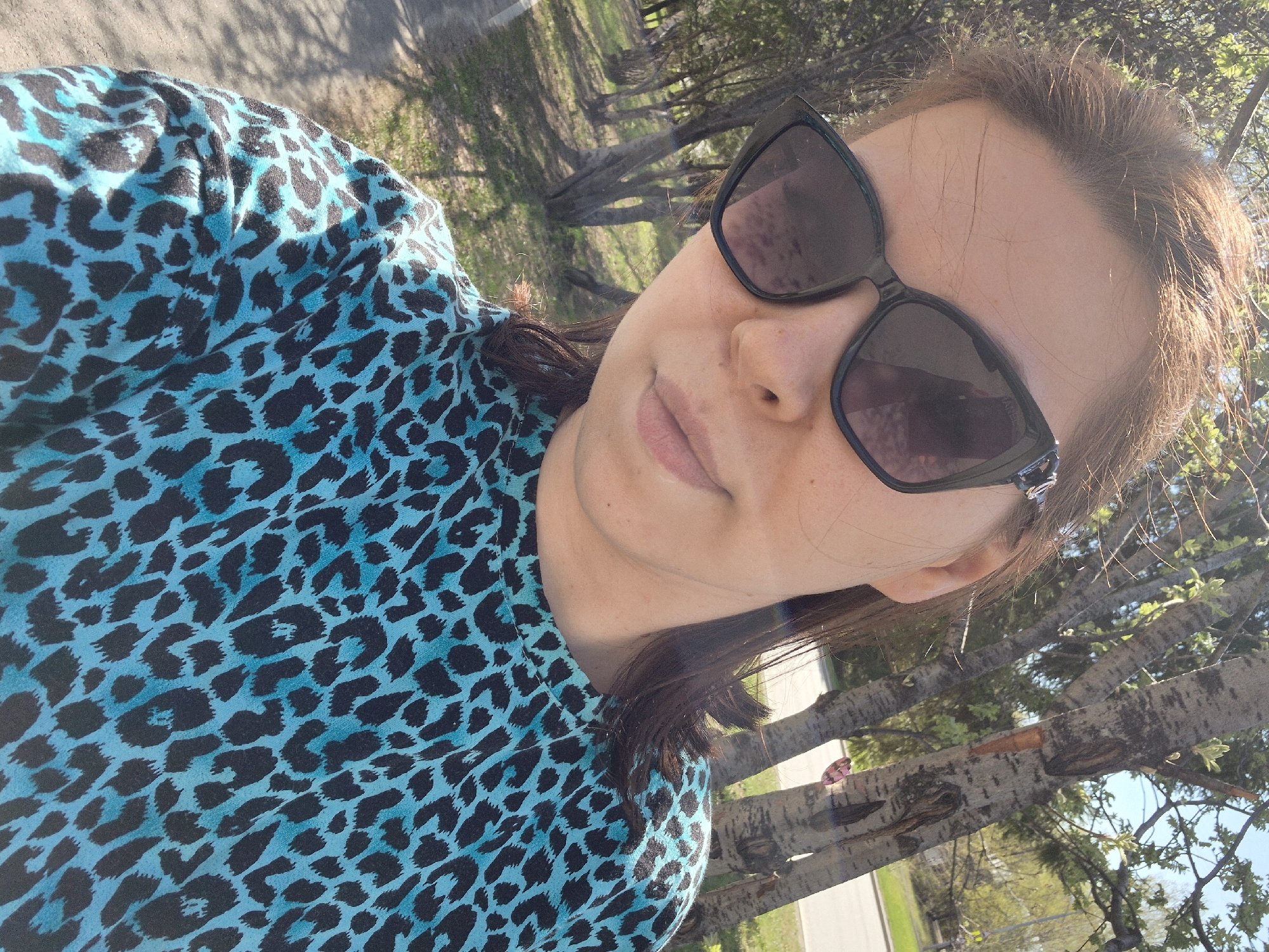 A selfie-style photograph shows a person wearing sunglasses and a blue animal-print top. The background features trees, a road, and possibly a park or green area. Specific landmarks that would locate the image within the city of Tobolsk, Russia, are not discernible in the image.