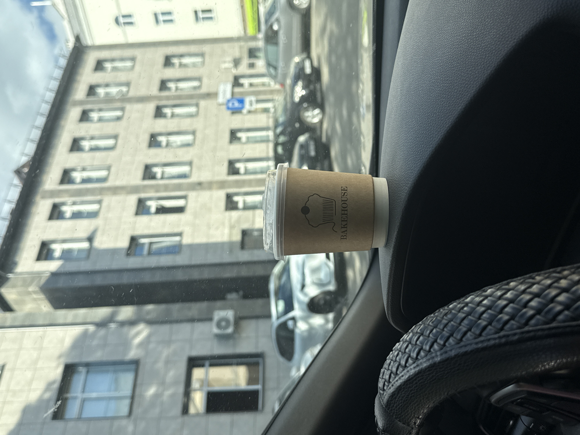 The image shows a coffee cup with the text "BAKEHOUSE" printed on it, placed on the dashboard of a car. The cup is brown and has a clear plastic lid. The background, visible through the car's windshield, depicts a building with multiple windows and an air conditioning unit. In the distance, several cars are parked on the street. The scene appears to be set in a city, likely Mahilioŭ, Belarus, during the daytime. The lighting suggests it's a sunny day, with shadows cast on the building. The overall impression is that of a casual moment, perhaps a commute or a brief stop.