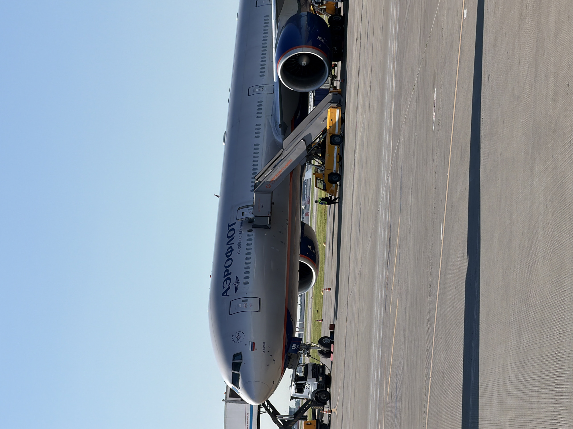 An Aeroflot Boeing 777-300ER aircraft, identified by the inscription "АЭРОФЛОТ" and "Российские авиалинии" on its fuselage, is parked on the tarmac at Sheremetyevo International Airport (SVO) in Khimki, Russia. The aircraft is a wide-body, twin-engine jet with the registration VQ-BGZ, named "И. Бунин". It is positioned at a gate, with a mobile passenger staircase connected to the forward left-side door. A yellow passenger stair truck is parked beneath the staircase. To the left of the aircraft, a white baggage tractor with the number "209" is visible, along with a catering truck to its left. Several orange traffic cones are placed on the tarmac around the aircraft. The sky above is clear and blue. The scene depicts typical airport ground operations.