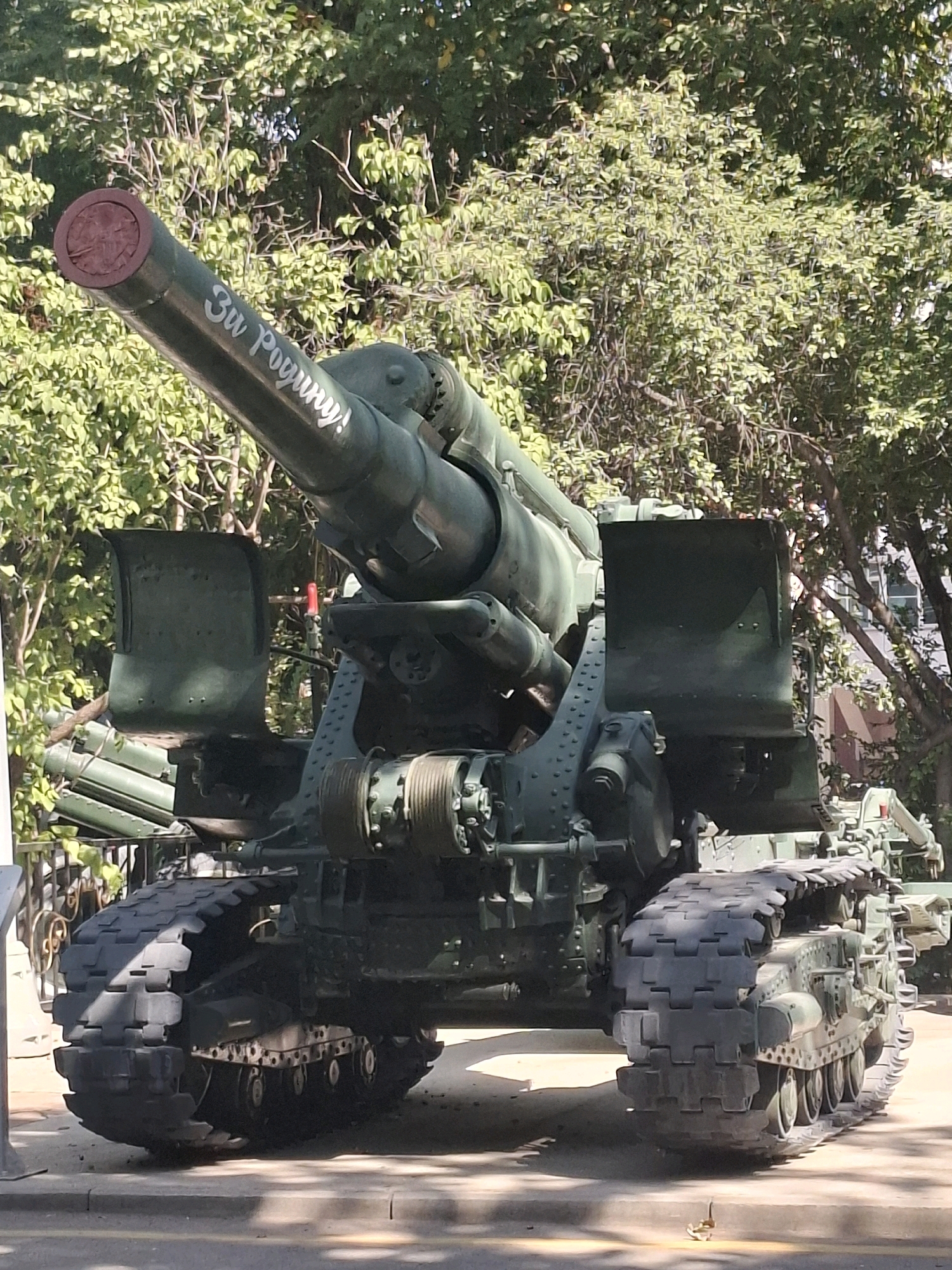 The image shows a large, green artillery piece with tracks, likely a self-propelled gun or a towed howitzer on a mobile platform, positioned outdoors. The cannon barrel is prominent, bearing white Cyrillic text that reads "За Родину!" (For the Motherland!). The artillery piece is situated in front of a dense, leafy green tree canopy, suggesting an outdoor setting with significant foliage. The lower part of the artillery piece rests on a paved surface, possibly a sidewalk or road, with shadows cast by the sun. To the left, a section of a metal fence or barrier is visible. The overall scene depicts a military artifact displayed publicly in a park-like or memorial environment. This specific artillery piece is located at the Panfilov Heroes Memorial Park in Almaty, Kazakhstan, where it serves as a monument. There are no people visible in the image.