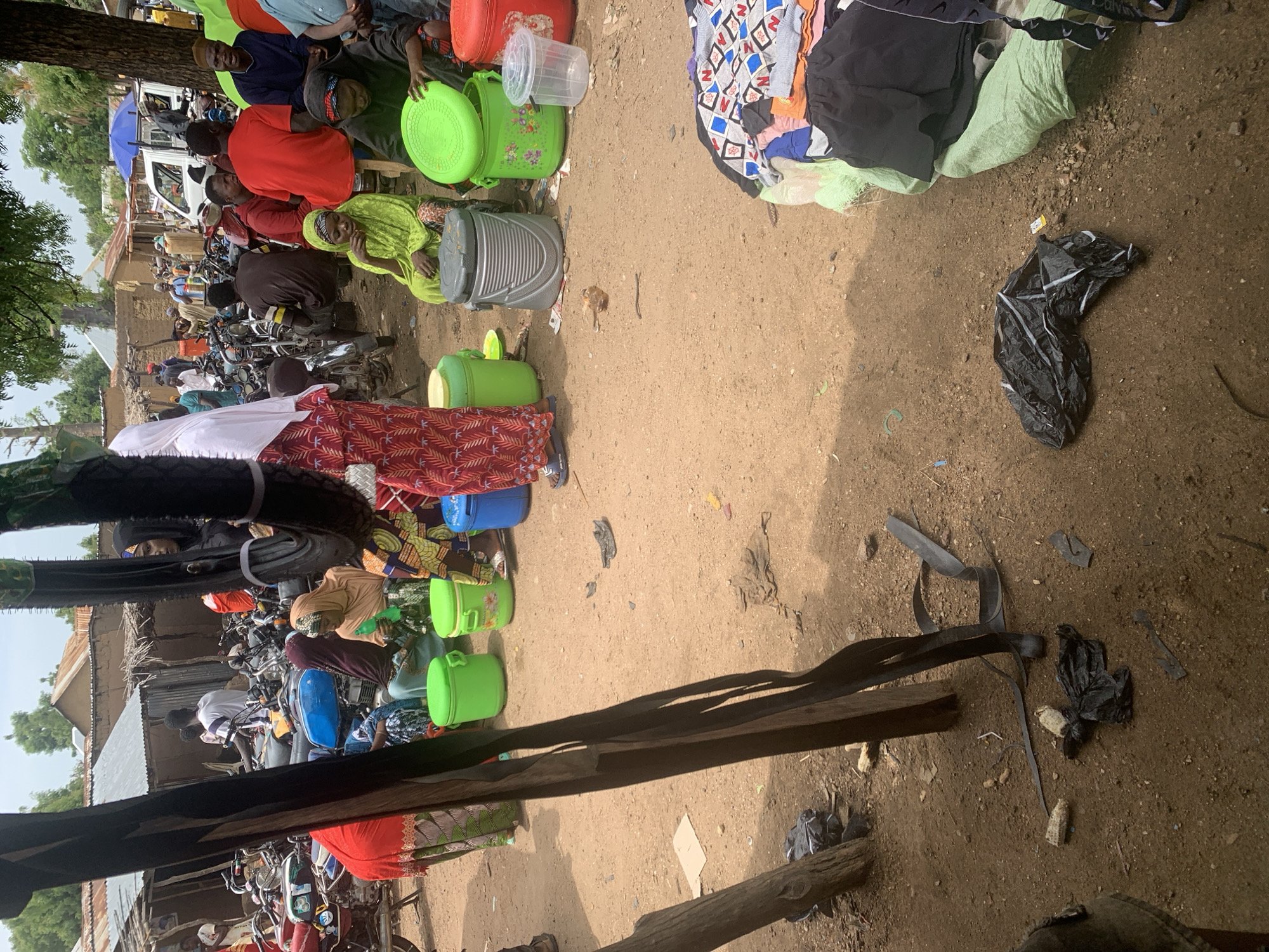 This media file depicts a bustling outdoor market scene in Nigeria, likely in the morning or early afternoon given the bright sunlight casting distinct shadows. The ground is a mixture of sandy soil and some scattered debris, suggesting a rural or semi-urban environment.
The primary subjects are a diverse group of people engaged in various activities within the market. Many individuals are dressed in colorful traditional clothing, with women often wearing headscarves and long dresses. Some people are seated, possibly vendors or shoppers taking a break, while others appear to be actively browsing or interacting. A significant number of motorcycles are parked closely together, indicating a common mode of transportation for market-goers and suggesting the market is a central hub.
A variety of objects are visible, predominantly containers. Large green plastic buckets and containers are scattered throughout the scene, some appearing new and others used. There are also a few clear plastic containers and a larger, grey, lidded bin. Piles of clothing, some folded and others loosely arranged, are also present, suggesting a clothing stall or a place where personal belongings are kept. A black plastic bag is visible on the ground, along with other assorted debris.
The overall atmosphere appears dynamic and busy. While specific emotions are hard to discern from still images, the crowded nature of the market and the presence of many people suggest a lively and active community gathering. The setting is characterized by makeshift stalls and the presence of trees and rudimentary structures in the background, further reinforcing the Nigerian market context. No visible text is identifiable within the image.