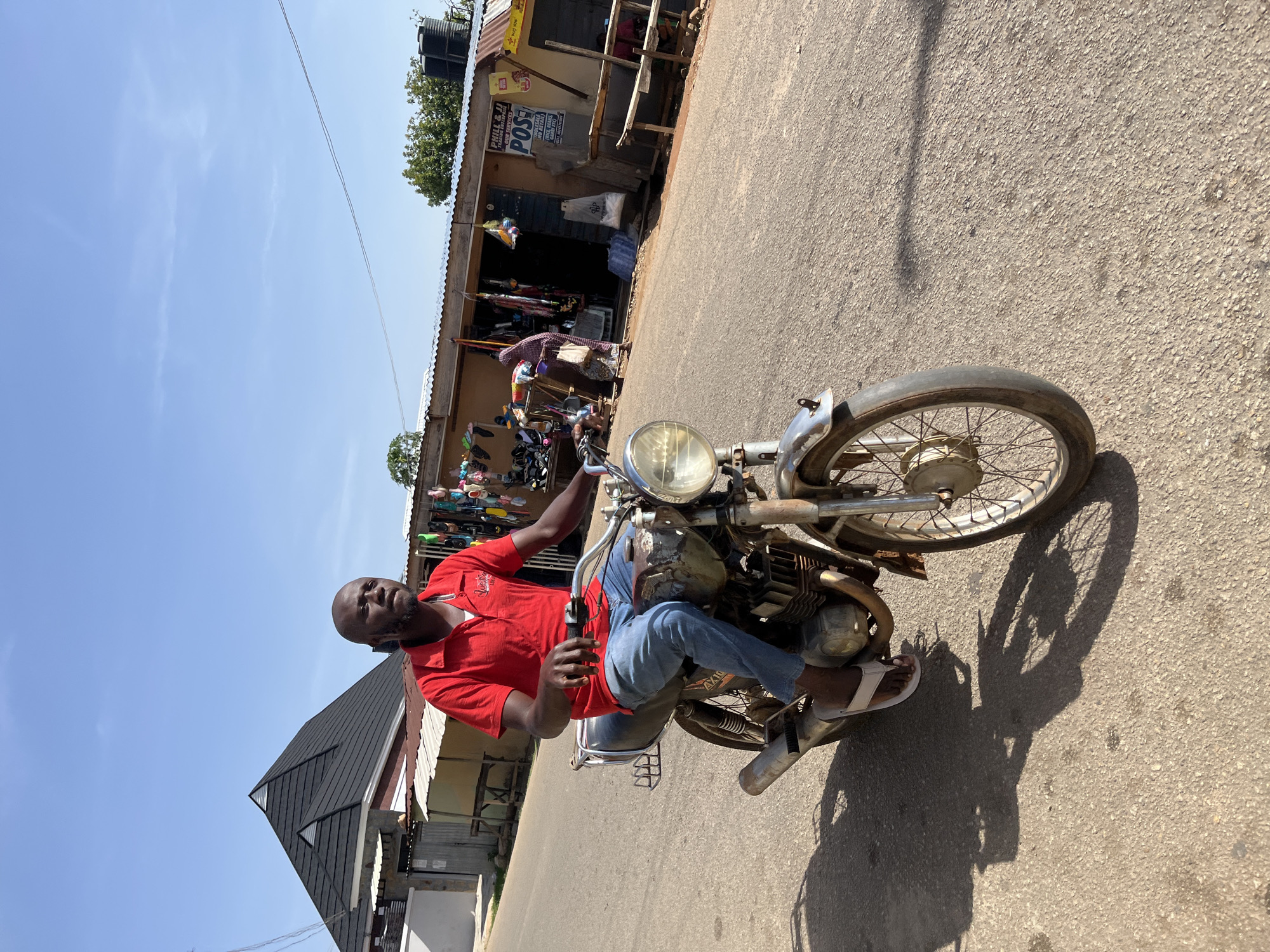 An eye-level shot captures a man seated on an older model motorcycle on a sunlit street in Jos, Nigeria. The man, who is bald, wears a red polo shirt, blue jeans, and white flip-flops, and looks directly at the viewer with a neutral expression. His hands are on the motorcycle's handlebars.
The motorcycle features a large, round headlight and a visible engine, showing signs of wear consistent with its age. It is positioned on an asphalt road, which appears somewhat worn. Strong, short shadows of the motorcycle and the man are cast on the pavement, indicating bright sunlight, likely around midday or early afternoon. The sky is clear and blue with very few clouds.
In the background, to the right, a row of small, makeshift-style shops or stalls lines the street. These structures have corrugated iron roofs, and various goods, including what appear to be shoes or slippers, are displayed outside. A black water tank is visible on the roof of one building. Several signs are visible on the stalls, with "PHAL & D" and "POS" clearly legible on two of them. To the left, a building with a dark, pitched roof is partially seen. The scene depicts a bustling, functional street environment in a town or city.