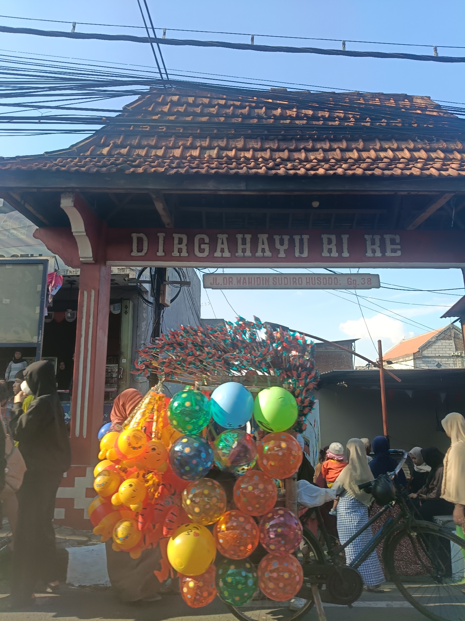 The image captures a street scene in Randuagung, Indonesia, under a clear blue sky. In the foreground, a vendor is selling balloons and pinwheels from a cart. The balloons are colorful, featuring various designs and characters, some shaped like smiley faces. Behind the balloons, a rack of red and green pinwheels is prominently displayed.
An archway with the Indonesian words "DIRGAHAYU RI KE" (congratulating the Republic of Indonesia on its anniversary) is visible above the scene. Below the arch, a street sign reads "JL. DR. WAHIDIN SUDIRO HUSODO: Gg. 38".
Several people are present in the image. On the left, a person in a black hijab walks away from the camera. Other individuals, mostly women wearing hijabs in various colors, are seen gathered near the vendor's cart and in the background. Some appear to be browsing the merchandise, while others are engaged in conversation. A bicycle with a basket is parked in the middle right of the frame.
The architecture suggests a typical Indonesian urban setting, with traditional tiled roofs and overhead electrical wires crisscrossing the sky. The overall atmosphere is lively, indicating a public space where goods are being sold and people are congregating. The presence of the celebratory banner suggests the photo was taken around Indonesia's Independence Day.