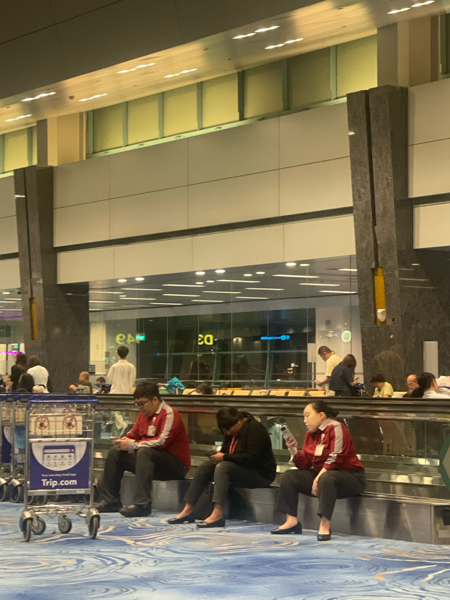 The image depicts an indoor environment that appears to be an airport or a large public transit hub, possibly Singapore Changi Airport. Several individuals are seated along a moving walkway or escalator railing. In the foreground, a luggage cart with the branding "Trip.com" is visible.
Two individuals in the middle ground are wearing red jackets with white accents and dark pants, possibly uniforms. One of them is looking at their phone. To their right, another person in dark clothing is also looking at their phone. Further to the right, a person in a similar red and white uniform is holding a bottle and looking at their phone. In the background, other people are visible, some standing and some seated, in a brightly lit area with reflective surfaces and architectural columns. The flooring has a distinctive blue and white swirling pattern.