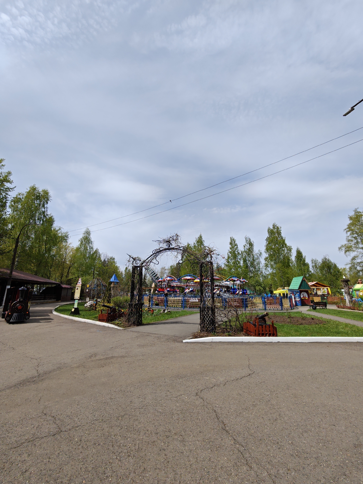 This image depicts an entrance to an amusement park in the city of Chaikovsky, Russia. The entrance features a decorative metal archway. Inside the park, there is a carousel ride and various themed structures, including small houses and a model train. The ground is paved with asphalt. The sky is overcast. Trees surround the park area.