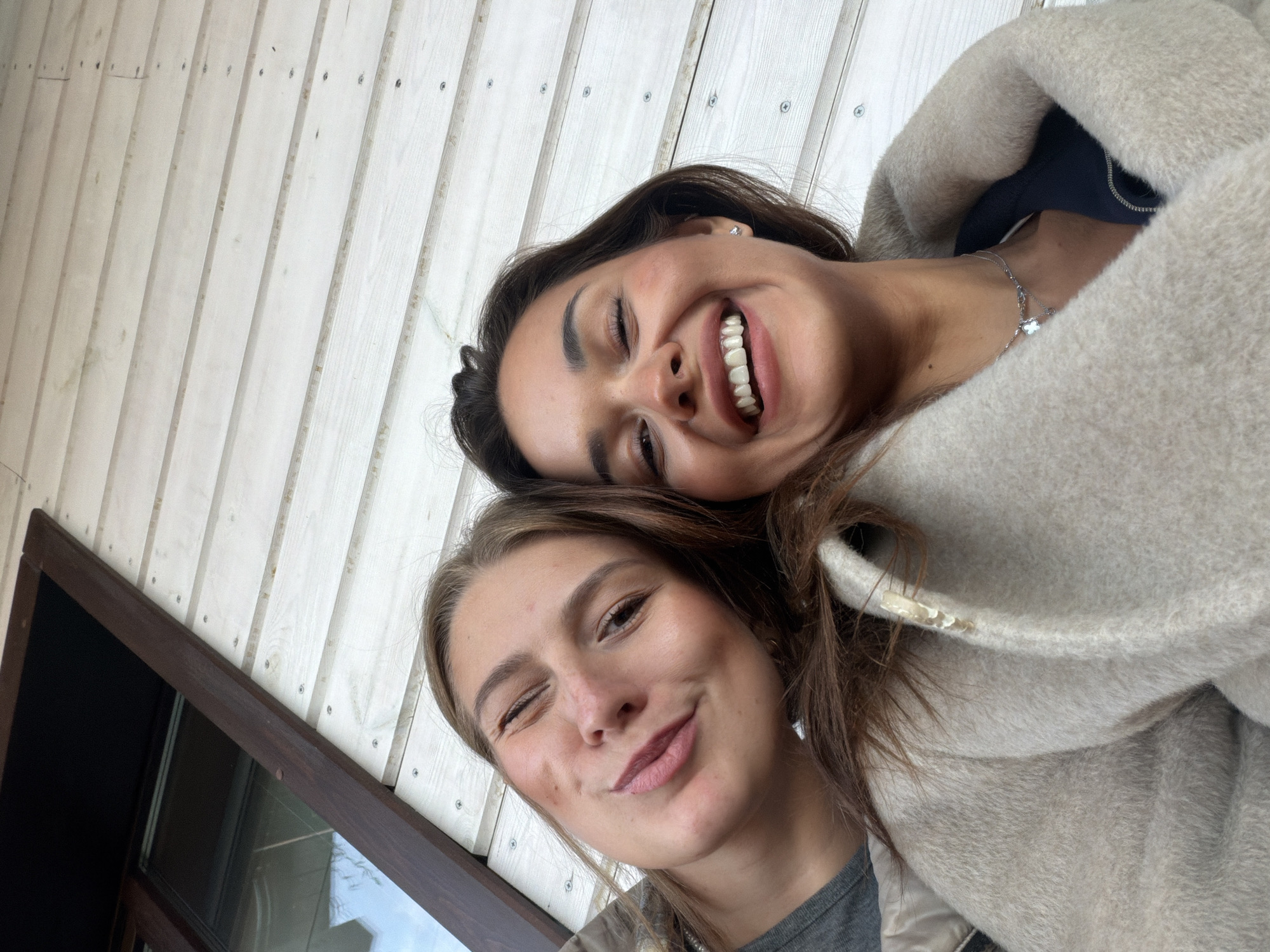 Two women are closely posed together, likely taking a selfie. The woman at the top is smiling broadly, exposing her teeth. She has dark hair and is wearing a light-colored coat or shawl. A silver necklace is visible. The woman below her is winking with one eye closed. She has light brown hair and is wearing a gray shirt. The background features light-colored, vertically aligned planks with visible screws. A dark brown frame is visible on the left, suggesting a window or architectural element. There is no verifiable information about the city of Shamoni, Russia.