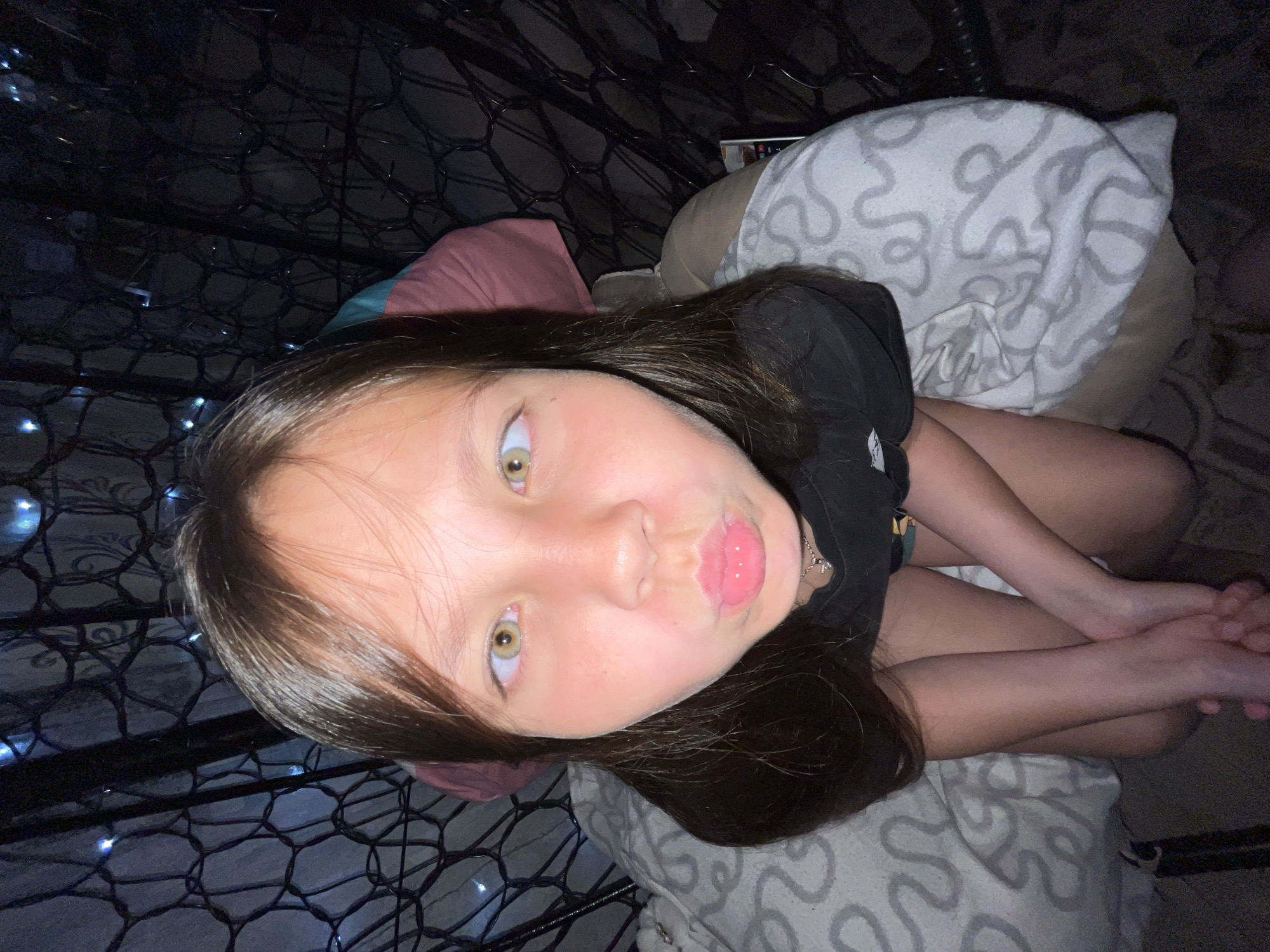 A young girl with long brown hair and fair skin is posing for a selfie in an indoor setting, potentially a bedroom. She is lying on a bed or cushion covered with a patterned white and gray fabric. The girl is wearing a black shirt and a silver necklace with a cross pendant. Her lips are pursed, and her eyes are directed towards the camera. In the background, there is a mesh or netting structure, possibly a part of a bed canopy or decorative element, adorned with small, lit bulbs. A pillow covered in a similar gray and white pattern is behind the girl. Part of her legs and hands are visible, as if she is lying down in a relaxed manner. The overall lighting is dim, creating a somewhat moody atmosphere. The location and city details are not discernible from the image.