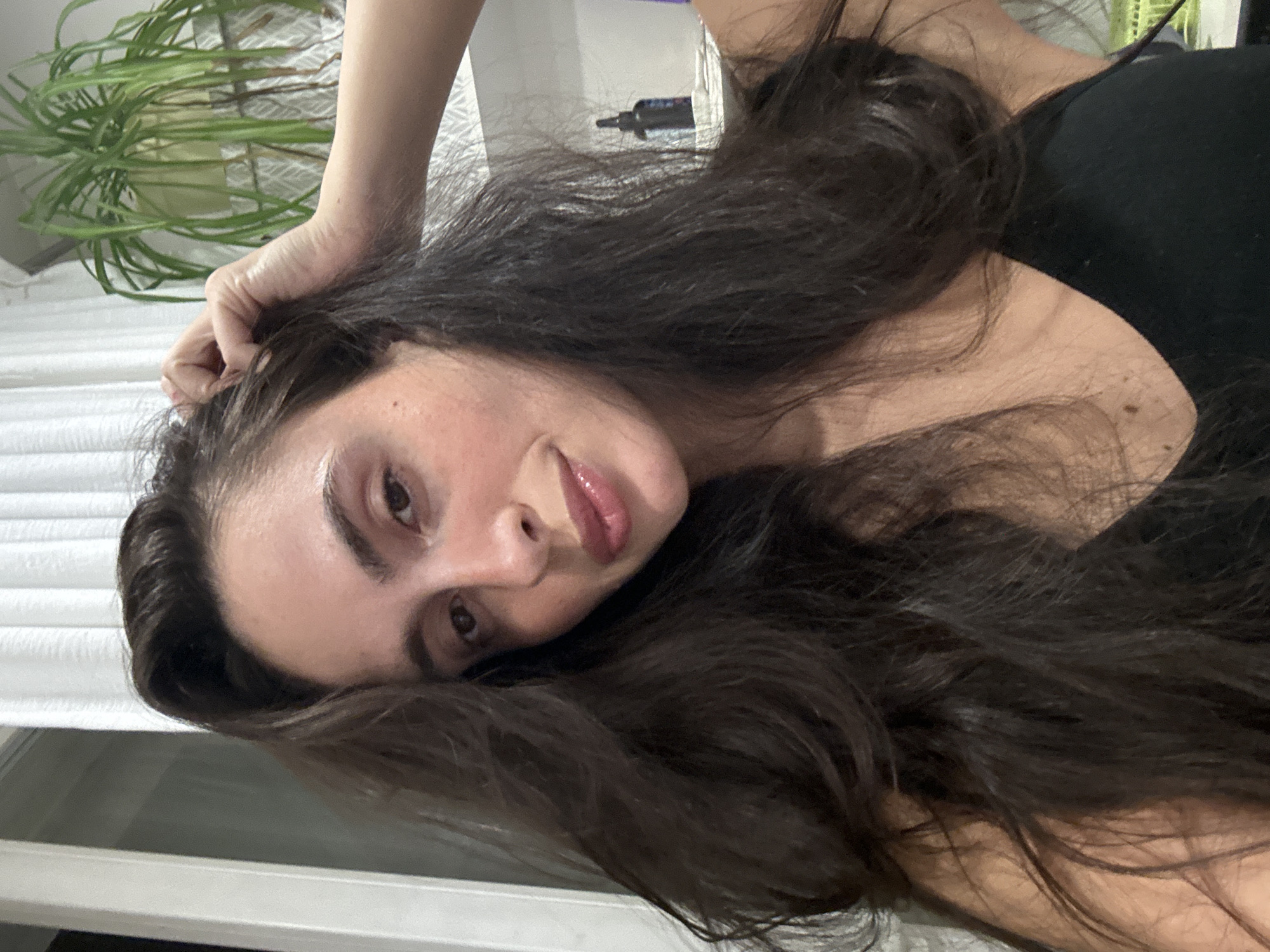 A woman with long, dark hair is lying down, looking towards the camera. Her left hand is near her head. She is wearing a black top. A potted green plant is visible in the upper left corner of the image. A bottle is located in the background. The background is a white, possibly indoor, surface. It is impossible to determine if this image was taken in Krasnoyarsk, Russia based solely on visual data.
