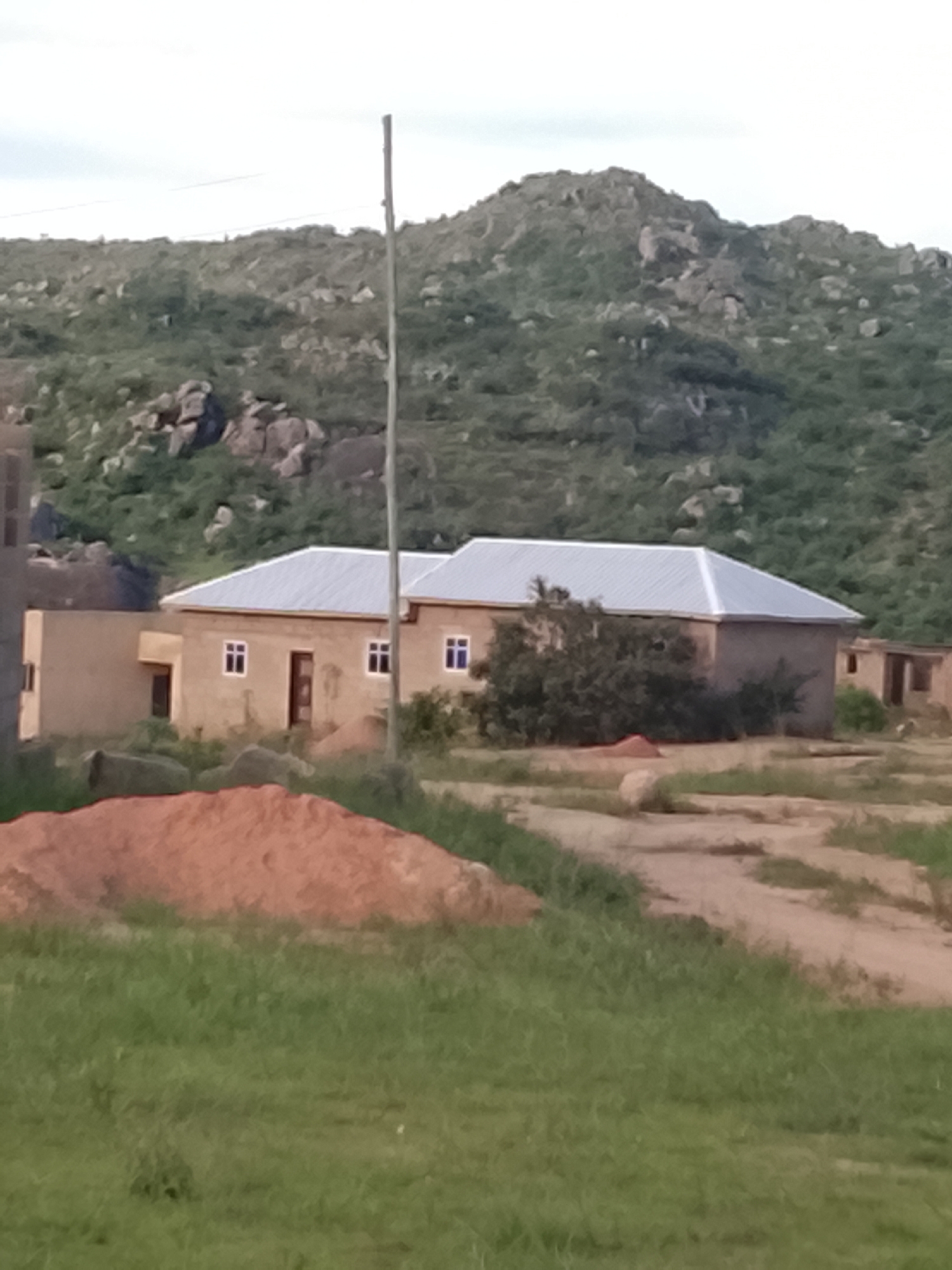 In the city of Barakin Acha, Nigeria, a long, gray, thin pole stands in the center of the frame, bisecting a light brown, single-story building with a silver, sloped roof. The building has several dark-framed windows and dark-colored doors. To the left of the main structure, another section of the building with a visible window and a dark door is present. Behind the building, a lush green hill dotted with large rocks and sparse vegetation rises. In the foreground, a pile of reddish-brown dirt is visible, with green grass surrounding it. The ground in front of the building appears to be a dirt path.