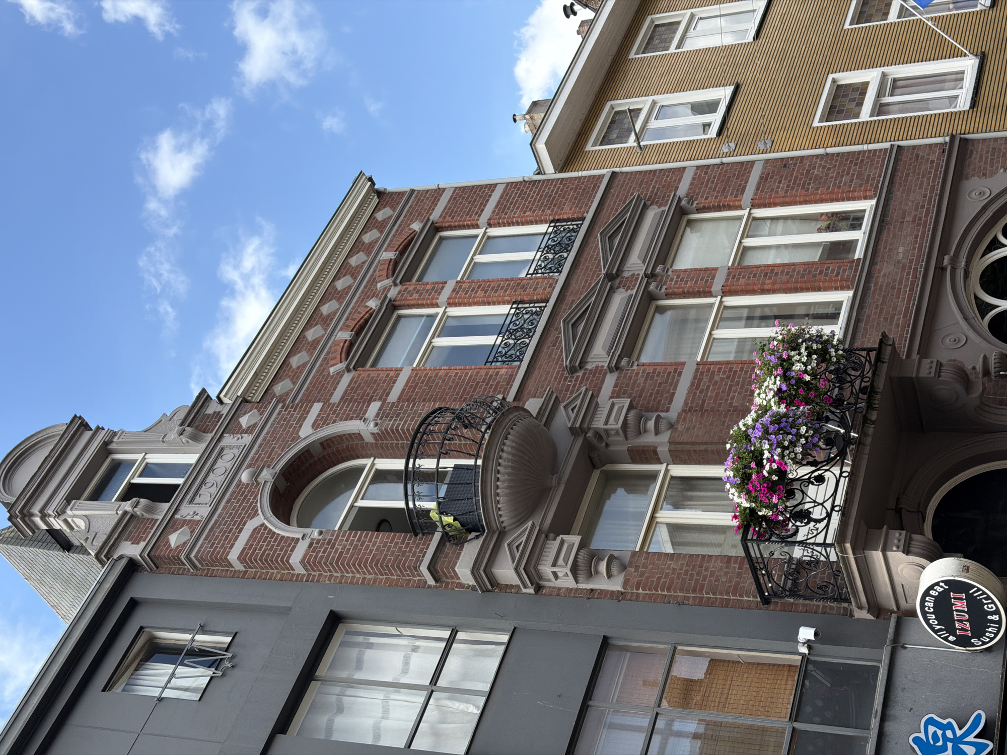 The image depicts a close-up view of the facades of several buildings in Roermond, Netherlands, under a bright blue sky with scattered white clouds. The central building is constructed of red brick with white trim, featuring ornate architectural details such as a decorative cornice, diamond-shaped accents, and a arched window with a black wrought-iron balcony above it. This balcony, like the one lower down on the same building, is adorned with a profusion of colorful flowers in shades of pink, purple, and white.
To the left of the brick building is a narrower, modern building with a dark gray facade and large, multi-paned windows. Some of these windows have white curtains, and one has a bamboo blind. A small white security camera is mounted on this facade. Below the windows of the gray building, a portion of a sign for "all you can eat IZUMI Sushi & Grill" is visible, featuring blue and white lettering on a black background.
To the right of the brick building is a building with a light yellow, horizontally ribbed facade. This building has several white-framed windows, some of which are smaller and appear to have decorative colored glass panes. A white flagpole extends from this building, though the flag it carries is only partially visible and appears to be blue and white. The overall impression is of an urban street scene with a mix of architectural styles, from historic brickwork to more contemporary designs.