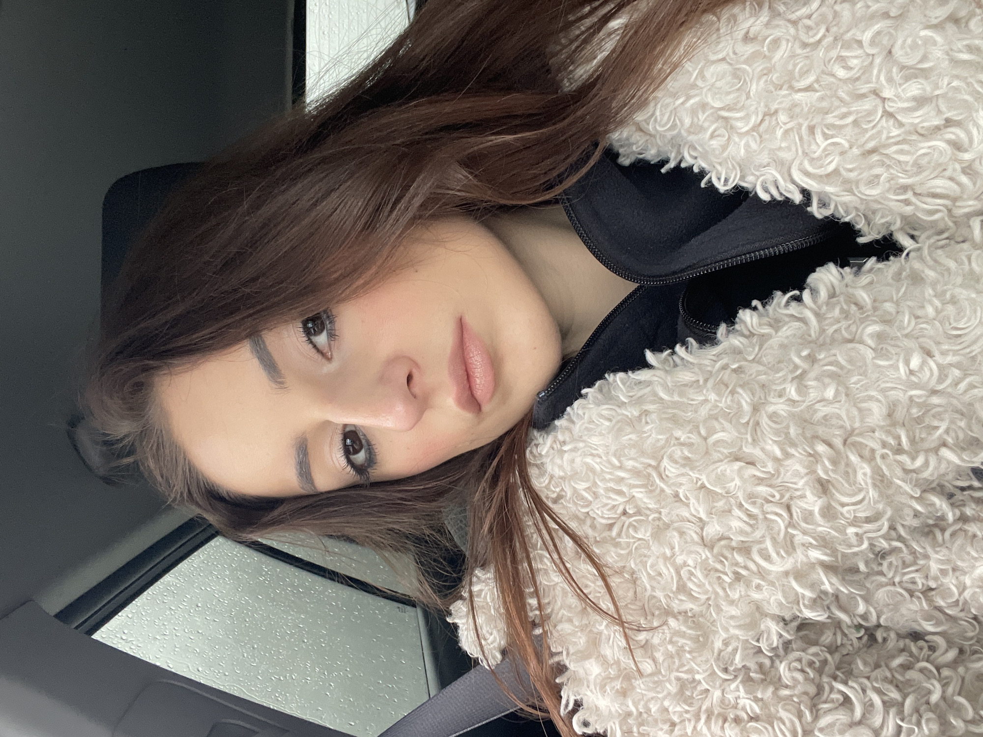 The image features a young woman positioned inside a car, captured in a close-up perspective. She has long, straight dark brown hair, which frames her face and falls over her shoulders. Her face is turned slightly towards the viewer, with a calm expression, and she is looking directly into the camera. Her makeup is subtle, featuring defined eyebrows, mascara, and light pink or nude lipstick.
She is wearing a layered outfit, consisting of a black zippered top or jacket underneath a thick, cream or light beige-colored coat. The coat has a distinct, curly, textured material, resembling sheepskin or a teddy bear coat. A dark-colored seatbelt strap is visible across her left shoulder and chest, confirming her location within a vehicle.
The setting is the interior of a car, indicated by the visible seatbelt and elements of the car's interior, such as a dark panel on the left and a lighter grey headliner or trim on the right. Through what appears to be a car window or sunroof, numerous raindrops are visible, suggesting it is raining outside during the daytime. No specific activity beyond posing for the photograph is depicted. There is no visible text in the image. This scene takes place within the context of Moscow, Russia.