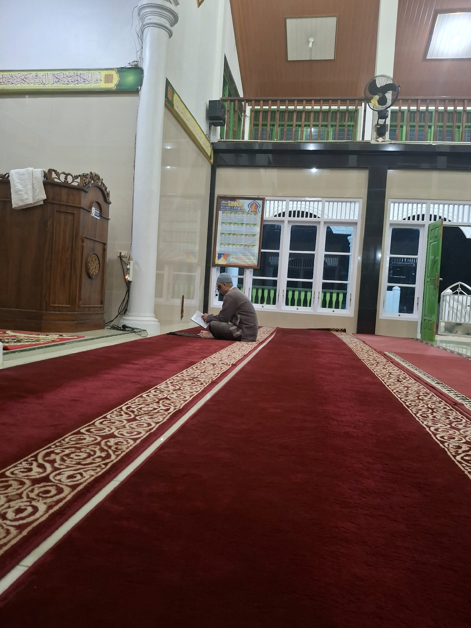 A single, middle-aged man wearing a light brown thobe and a light blue skullcap is seated on a red carpet in a mosque, reading a book. He is positioned to the right of the frame, facing left. The mosque features a large wooden podium on the left, with white towels draped over it. A white pillar is visible behind the podium. The walls are light-colored, with green and gold Arabic calligraphy running along the top. The floor is covered by a plush red carpet with a decorative border. In the background, there are large windows with white frames and green shutters. A black oscillating fan is mounted on a shelf above the windows. The scene is illuminated by artificial light. The location is identified as a mosque in Yogyakarta, Indonesia.