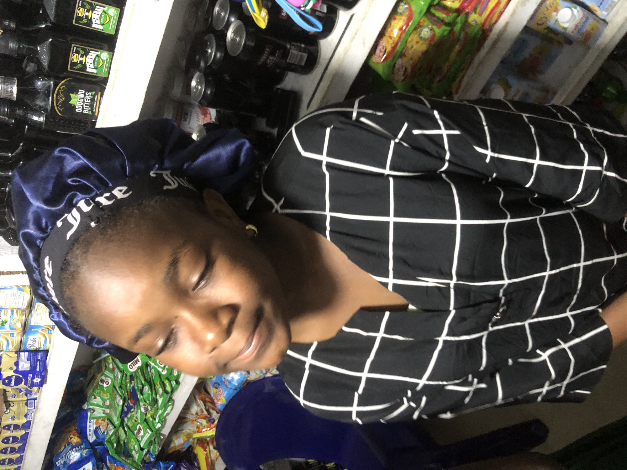 A young Black girl, wearing a black and white checkered shirt and a blue satin bonnet with white lettering, is pictured in what appears to be a small convenience store in Ado, Nigeria. She is looking downwards with her eyes closed and a slight smile on her face. The shelves behind her are stocked with various products, including what look like juice boxes and bottles of bitters. The lighting is dim, suggesting it might be late in the day or the store has limited artificial light.