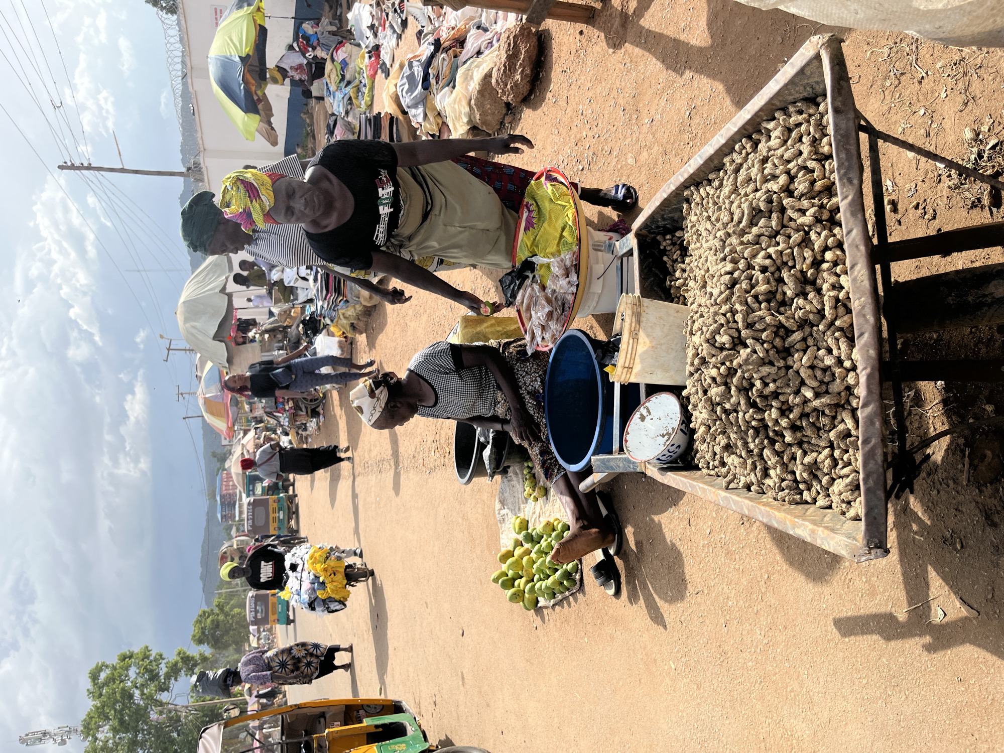 The image depicts an outdoor marketplace in Rafin Par, Nigeria, under a cloudy sky. A dirt road forms the thoroughfare, with several individuals engaged in commerce. In the foreground, a large cart is filled with peanuts in their shells. To the left, a woman sits on the ground beside a pile of green mangoes and a large blue basin. Further back, a vendor stands behind a tray holding small packages and a colorful cloth. Other people are seen walking and interacting within the market, with some pushing carts and others attending to stalls. Various goods, including clothing and other merchandise, are visible on display and piled on the ground. The presence of a cell tower and utility poles suggests an urban or semi-urban setting. The overall scene is bustling and active, characteristic of a local market environment.