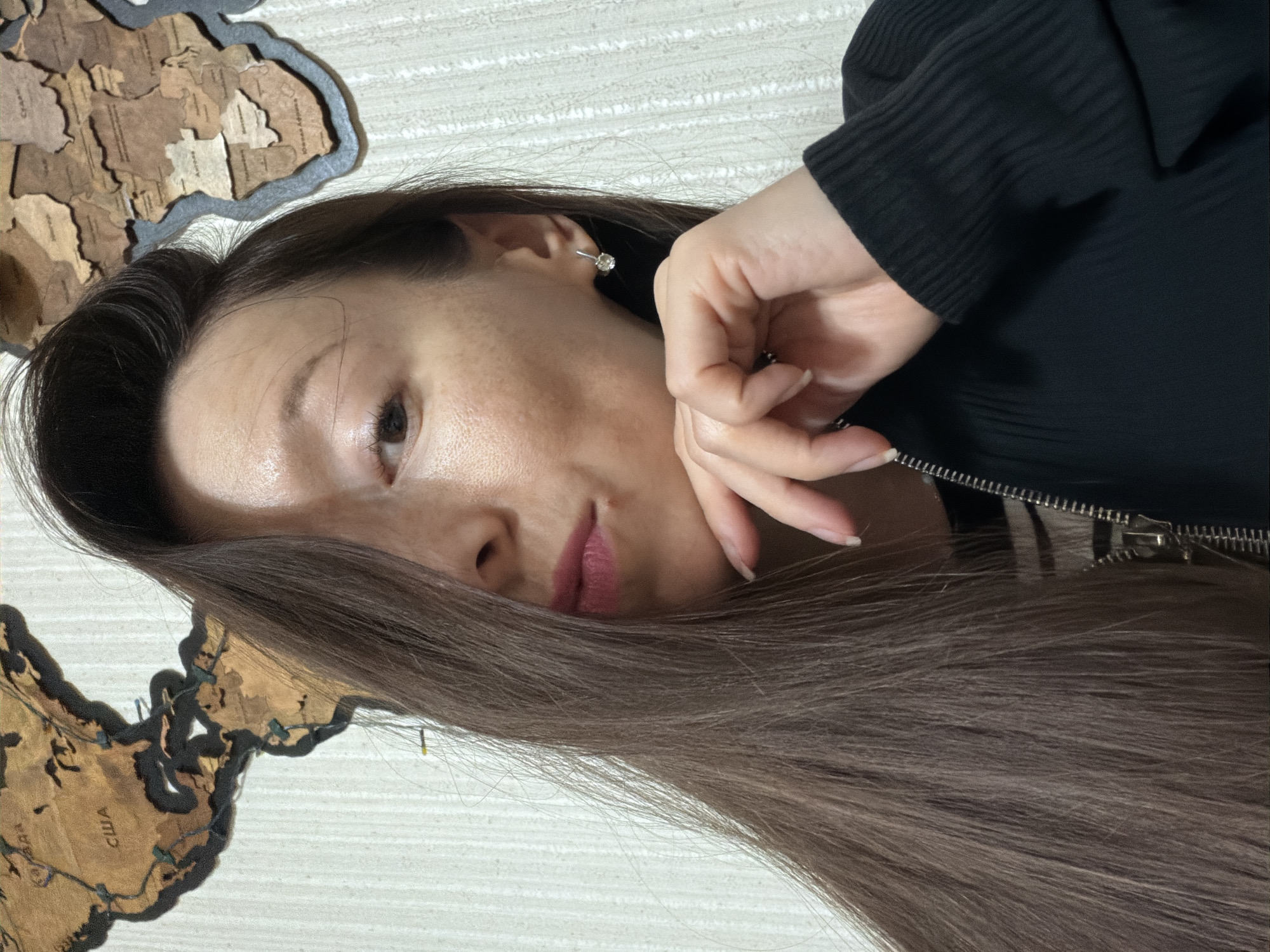 A young woman with long, dark brown hair covering half of her face is shown from the chest up. She is wearing a black zip-up jacket over a striped top and a small stud earring in her visible ear. Her right hand is resting on her chin, and her lips are slightly parted. The background consists of a light-colored textured wall and a wooden world map decoration mounted above and behind her, which appears to depict North America and Africa. No specific location in the city of Raevka, Russia is discernible.