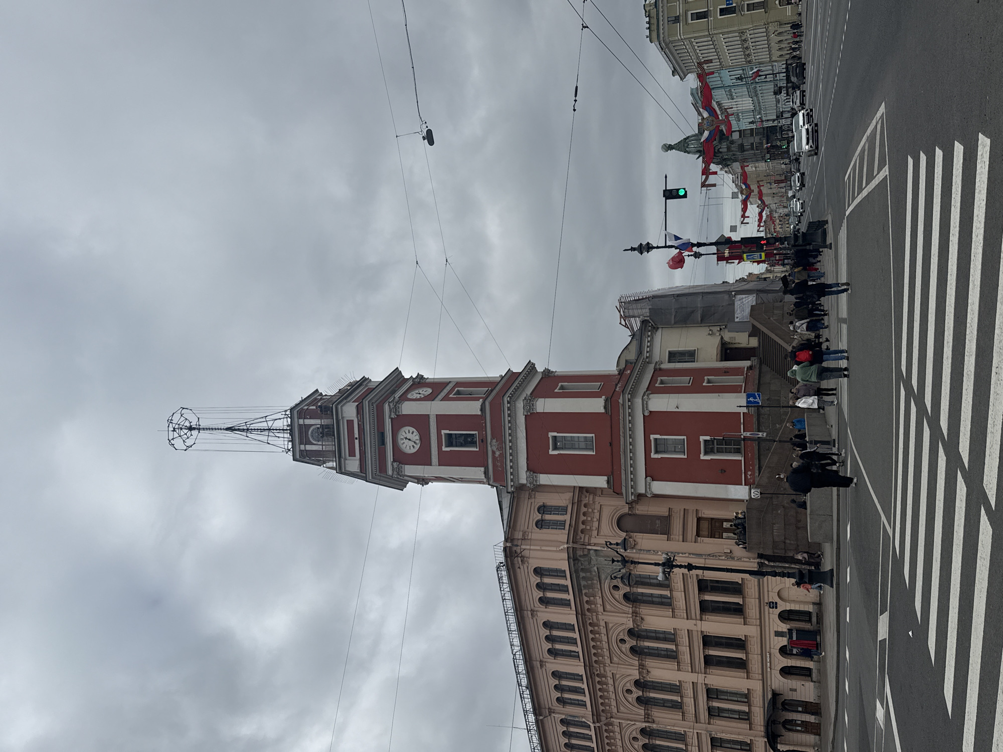The image depicts a street scene in Saint Petersburg, Russia, dominated by the Rostral Columns, specifically the one with red and white coloration. It's a daytime shot under an overcast sky. A number of pedestrians are shown using a crosswalk, with vehicles visible further down the street, which is adorned with flags (possibly Russian). The architecture is characterized by classical buildings with arched windows. Electrical wires are strung across the sky.