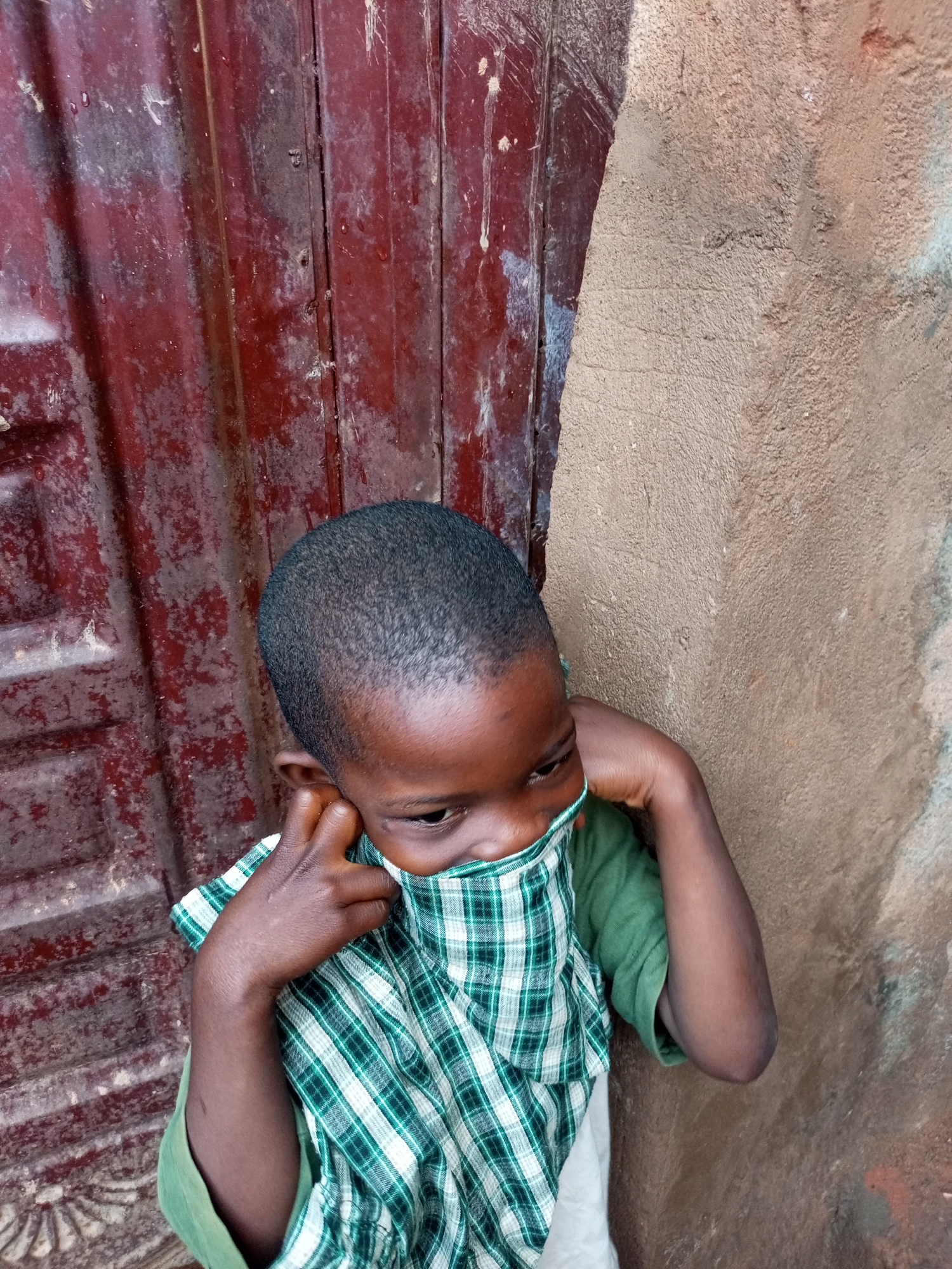 A young child with dark skin and short, dark hair is shown from the chest up. They are wearing a green t-shirt and a green and white plaid garment, possibly a shirt or scarf, pulled up to cover their nose and mouth. The child's head is turned to the right, and their right hand is touching their ear, while their left arm is bent with their hand near their face. The background consists of a textured, reddish-brown corrugated metal surface on the left and a rough, light brown stucco wall on the right. The image appears to be taken outdoors. The context of Tempure, Nigeria, is not visually discernible from the image itself.