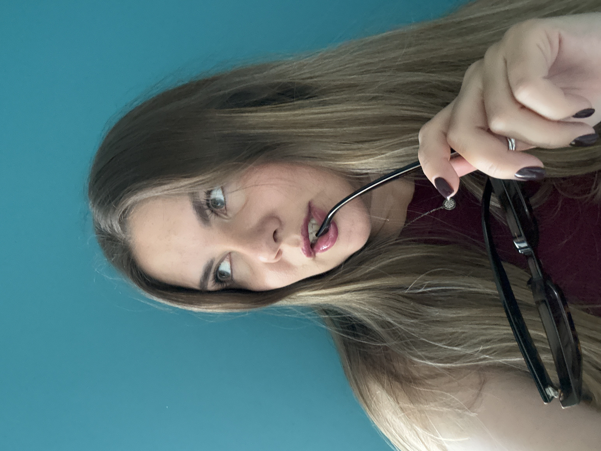 A woman with long, blonde hair holds a pair of black-framed glasses to her lip while looking at the camera. She has light skin, light eyes, and dark nail polish. She is wearing a maroon shirt and a silver necklace with a small pendant. The background is a solid turquoise color. The location is not identifiable as Izhevsk, Russia.