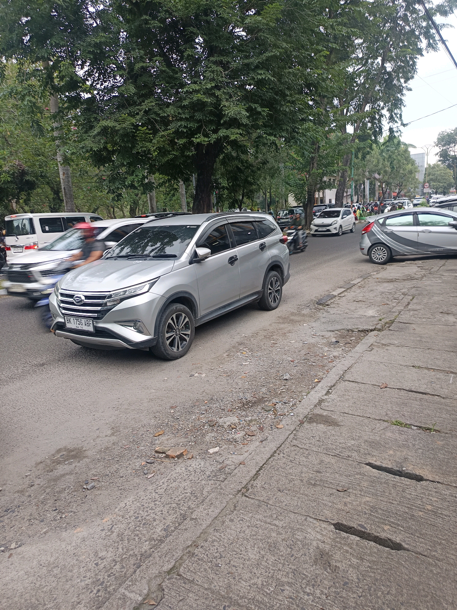 A street scene in the city of Medan, Indonesia, features several vehicles, including a silver SUV in the foreground, a white van, and other cars and motorcycles on the road. Trees line the street, with buildings visible in the background. The road surface is asphalt, with a concrete sidewalk on the right side of the image. The scene appears to be a busy urban environment with moderate traffic.
