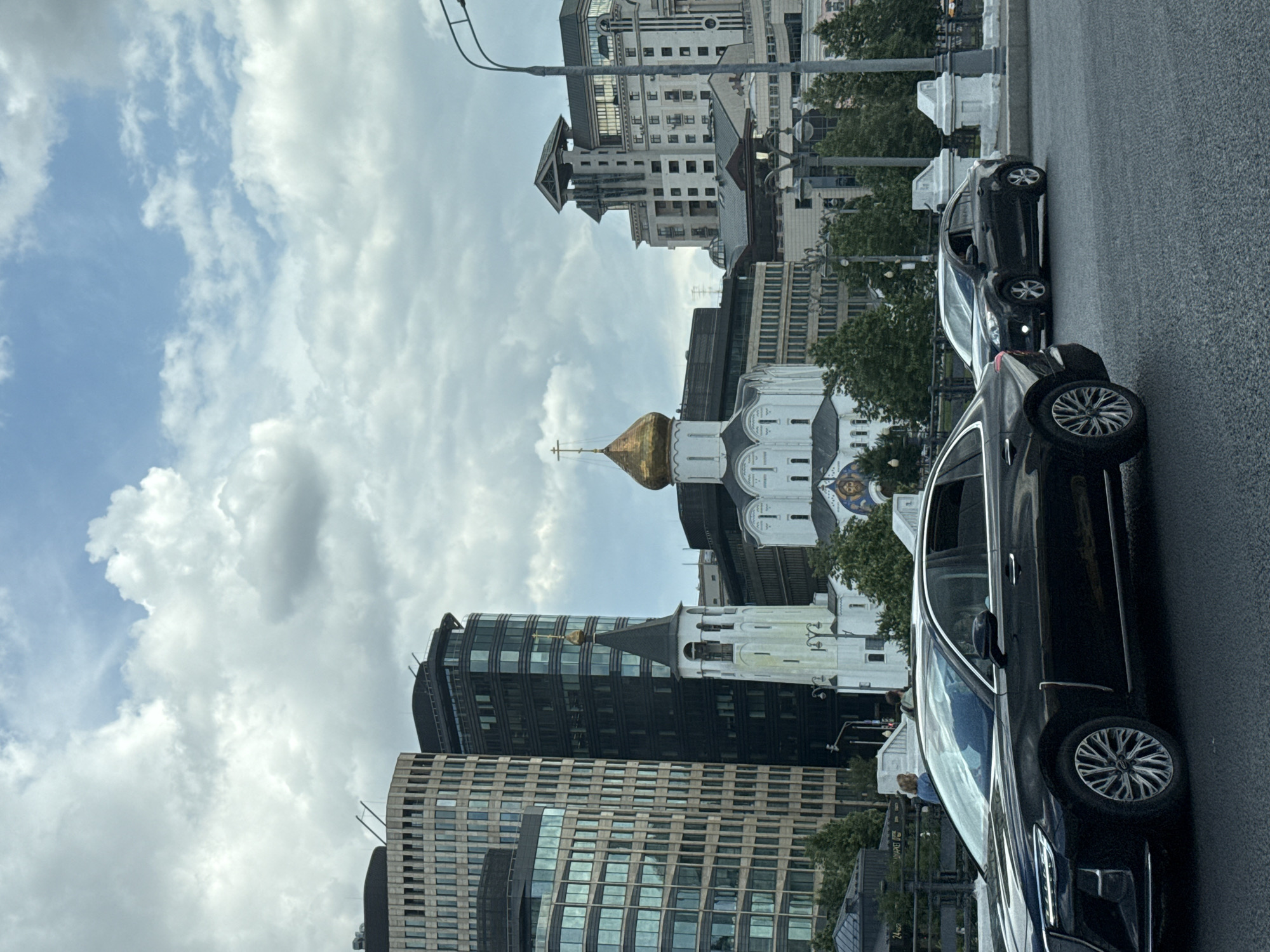 The image shows a street scene in Moscow, Russia, with several modern buildings and a historical church. In the foreground, a black sedan is driving on a paved road. Behind it, another dark-colored sedan is also visible. A bridge railing runs along the side of the road, with a few people walking on the other side.
In the background, a white church with a golden dome and a tall bell tower stands out amongst several tall, contemporary buildings. The church has a cross on its dome and a mural above its entrance. The buildings surrounding the church are a mix of styles, with some featuring glass facades and others having a more traditional architectural design. Trees and streetlights are also visible in the scene. The sky is cloudy, with patches of blue sky showing through. The overall impression is of a city where modern architecture coexists with historical landmarks.