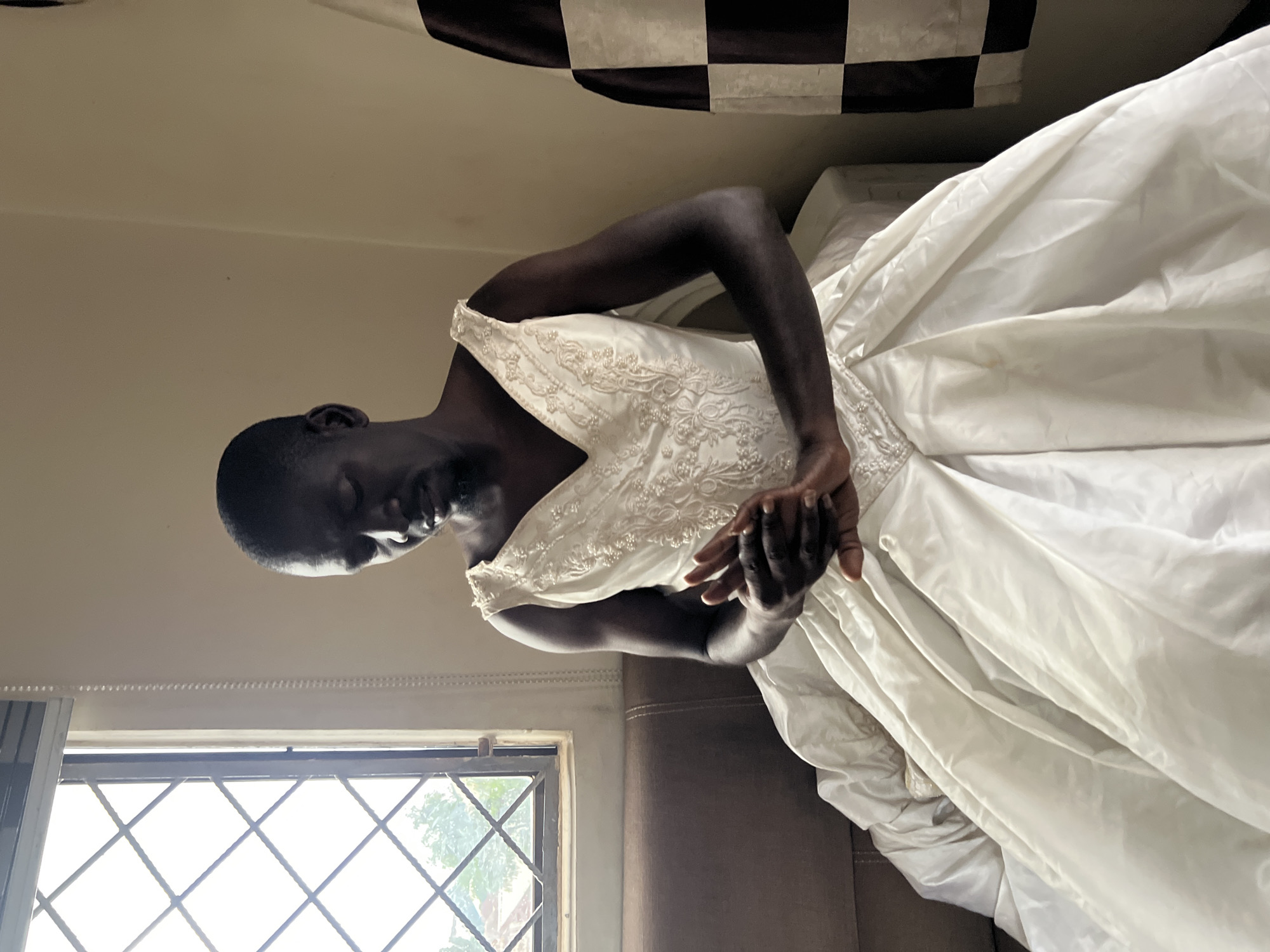 A dark-skinned man wearing a white wedding dress is sitting on a brown couch with his eyes closed and hands clasped. He has a short haircut and is looking down. The wedding dress is full-skirted and appears to be made of satin with beaded detailing on the bodice. To his left, a window with diamond-shaped panes looks out onto trees and buildings. To his right, a brown and white checkered curtain hangs. The scene is indoors, with a light-colored wall behind the man. The overall lighting is soft.