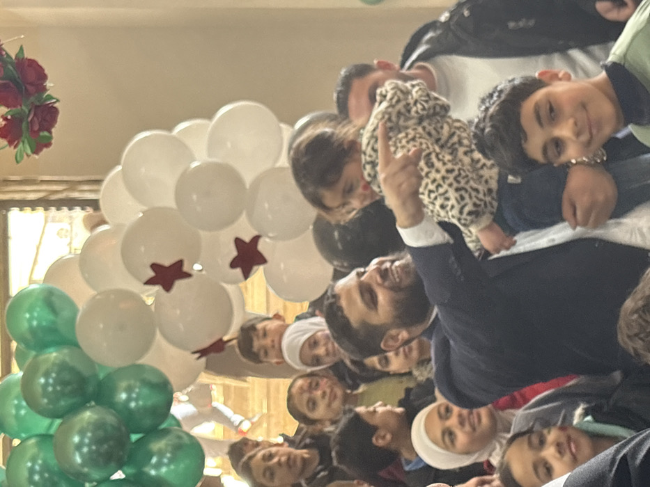 An indoor scene in Kafr Batna, Syria, depicts a group of individuals, predominantly children, gathered under a balloon arch. A bearded adult male, positioned centrally, is holding a small child wearing a leopard-print jacket. The man is smiling and pointing with his right hand towards an unseen area to the viewer's right. The child in his arms is also looking in that direction, with a faint red mark on her left cheek. Several other children of varying ages are visible around them, some smiling, and some wearing headscarves. In the background, an arrangement of green and white balloons forms an arc, with some white balloons featuring red star decals. A red floral decoration is partially visible in the upper right quadrant of the image. Other adults are partially visible in the background. The lighting suggests an interior space with artificial illumination.