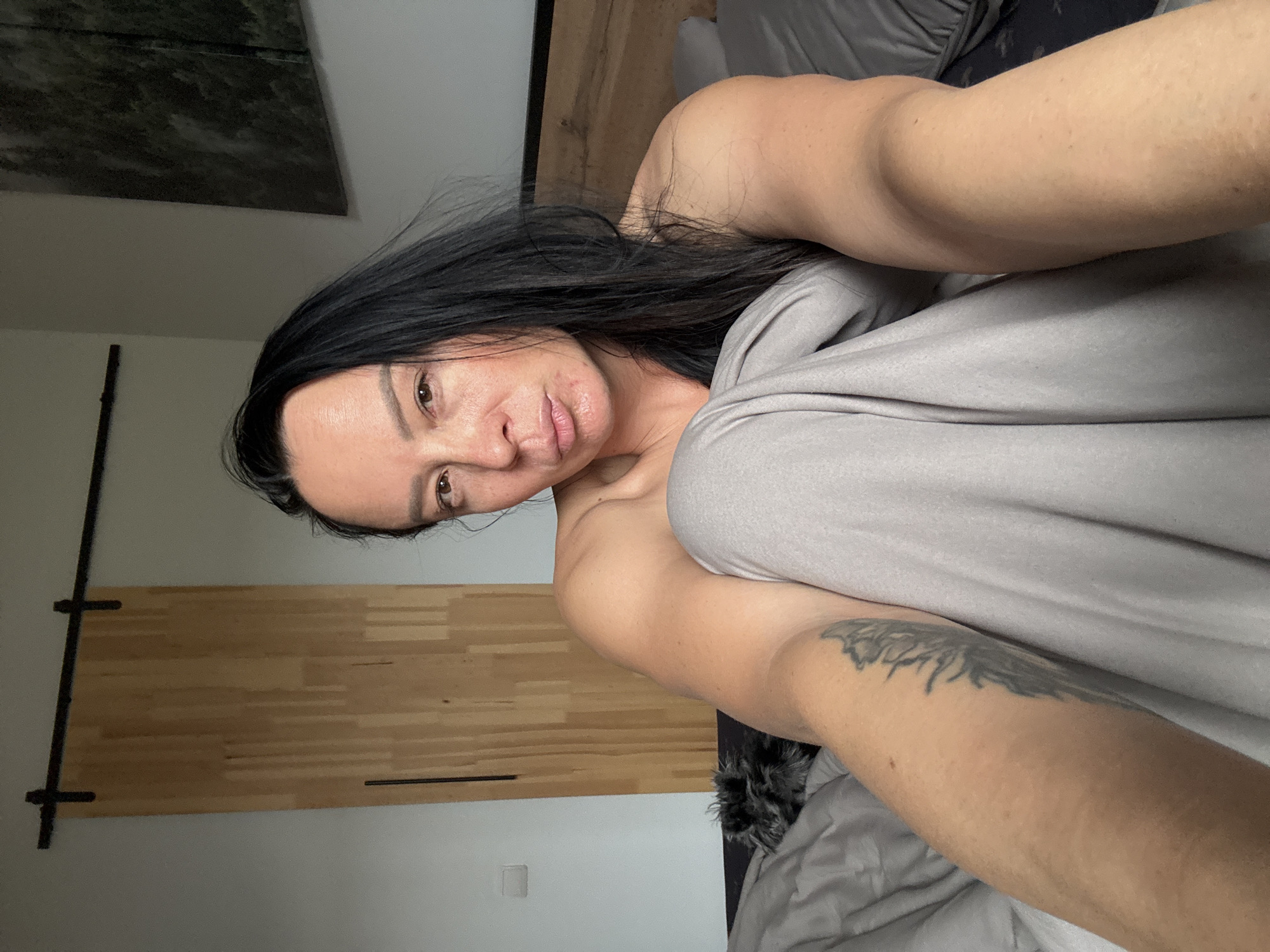 A woman with dark hair, partially covered by a gray blanket, lies in bed. A wooden headboard is visible behind her. A piece of art hangs on the wall. The location is most likely not Chaikovsky, Russia as no location details are visible.