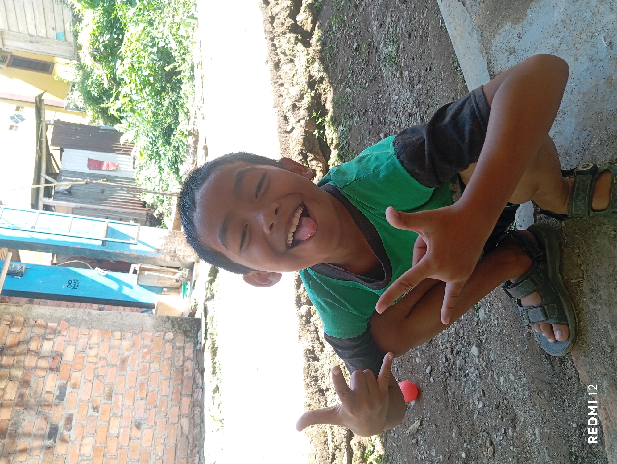 A young boy, estimated to be between 6-8 years old, is captured in a medium shot, crouching outdoors. He wears a green t-shirt with dark sleeves and grey sandals. The boy is making a "rock on" hand gesture with his left hand, thumb and pinky finger extended, and his index and middle fingers curled down, while his right hand is also in a similar gesture, but with the index finger pointed upwards and the middle finger extended outwards, and his thumb held in place with his ring and pinky fingers. His tongue is sticking out, and his mouth is wide open, suggesting a playful or energetic demeanor.
The background consists of a gravelly ground, a raised concrete edge, and a brick wall on the left. In the distance, there are structures, including corrugated metal siding and a yellow building. Green foliage is visible behind the structures, suggesting a semi-urban or rural setting. The lighting indicates a sunny day, with harsh shadows cast on the ground. The presence of "REDMI 12" watermark suggests the image was taken with a Xiaomi Redmi 12 smartphone. The location is identified as Pagar Alam, Indonesia.